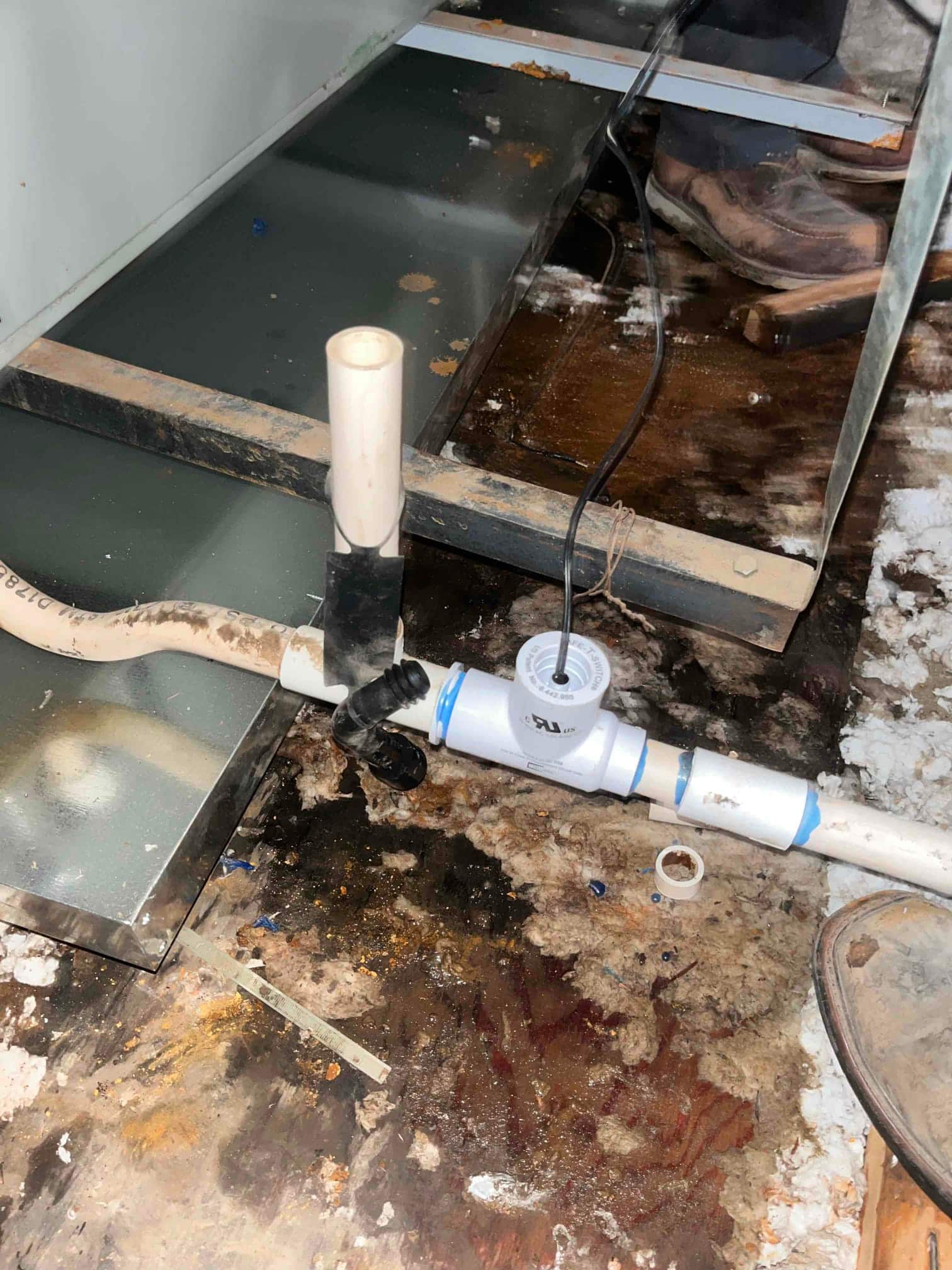 Condensate drain line with tee and cleanout cap beside the air handler, with surrounding wet floor and insulation.