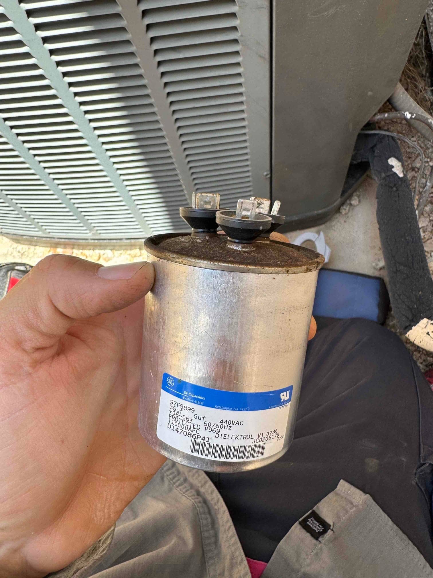 Cylindrical run capacitor held near an outdoor condenser, showing two spade terminals and rust discoloration on the top.