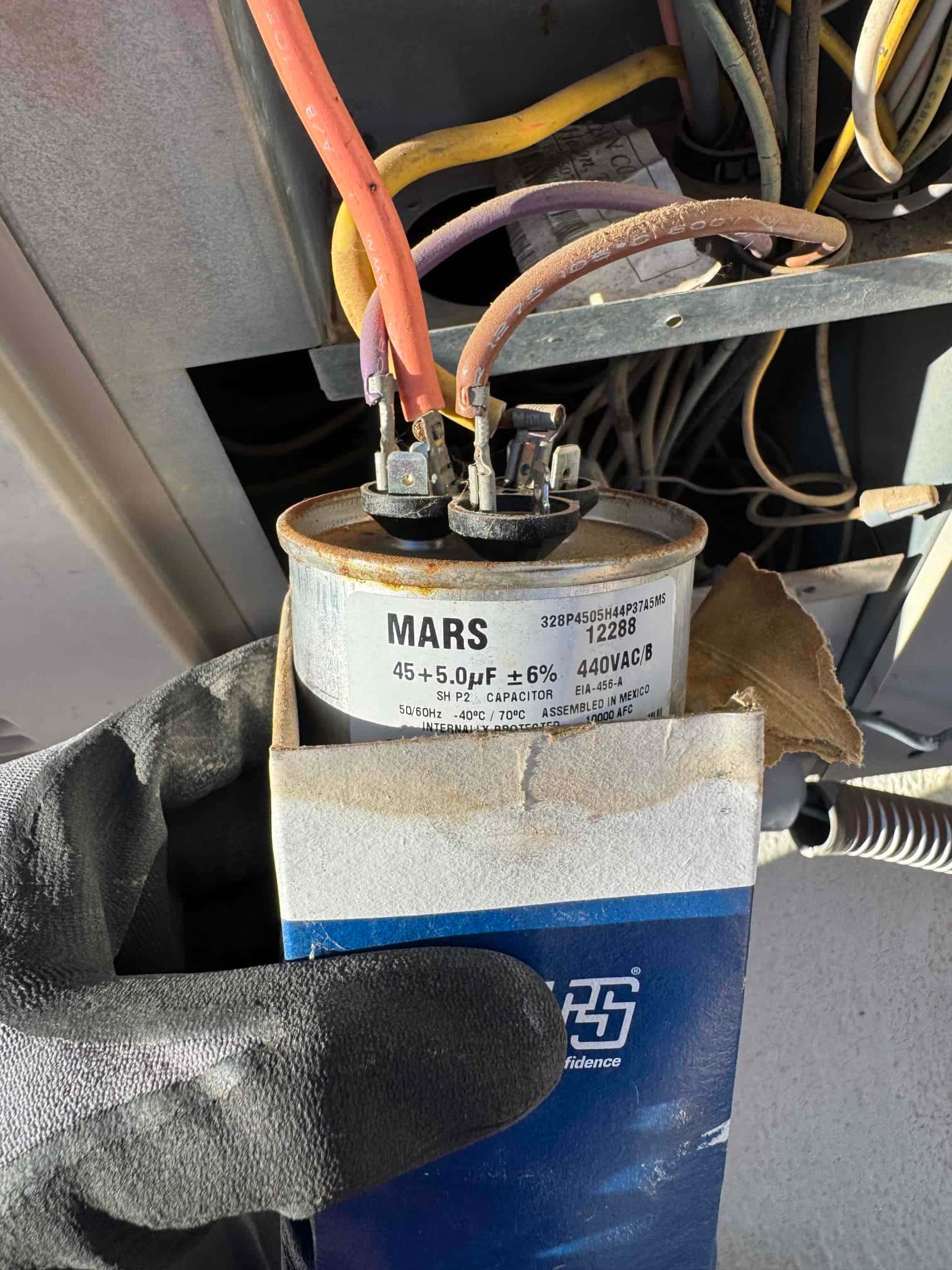 Dual-run capacitor labeled 45+5.0 μF ±6% 440VAC, held in a gloved hand with wires attached to its terminals.