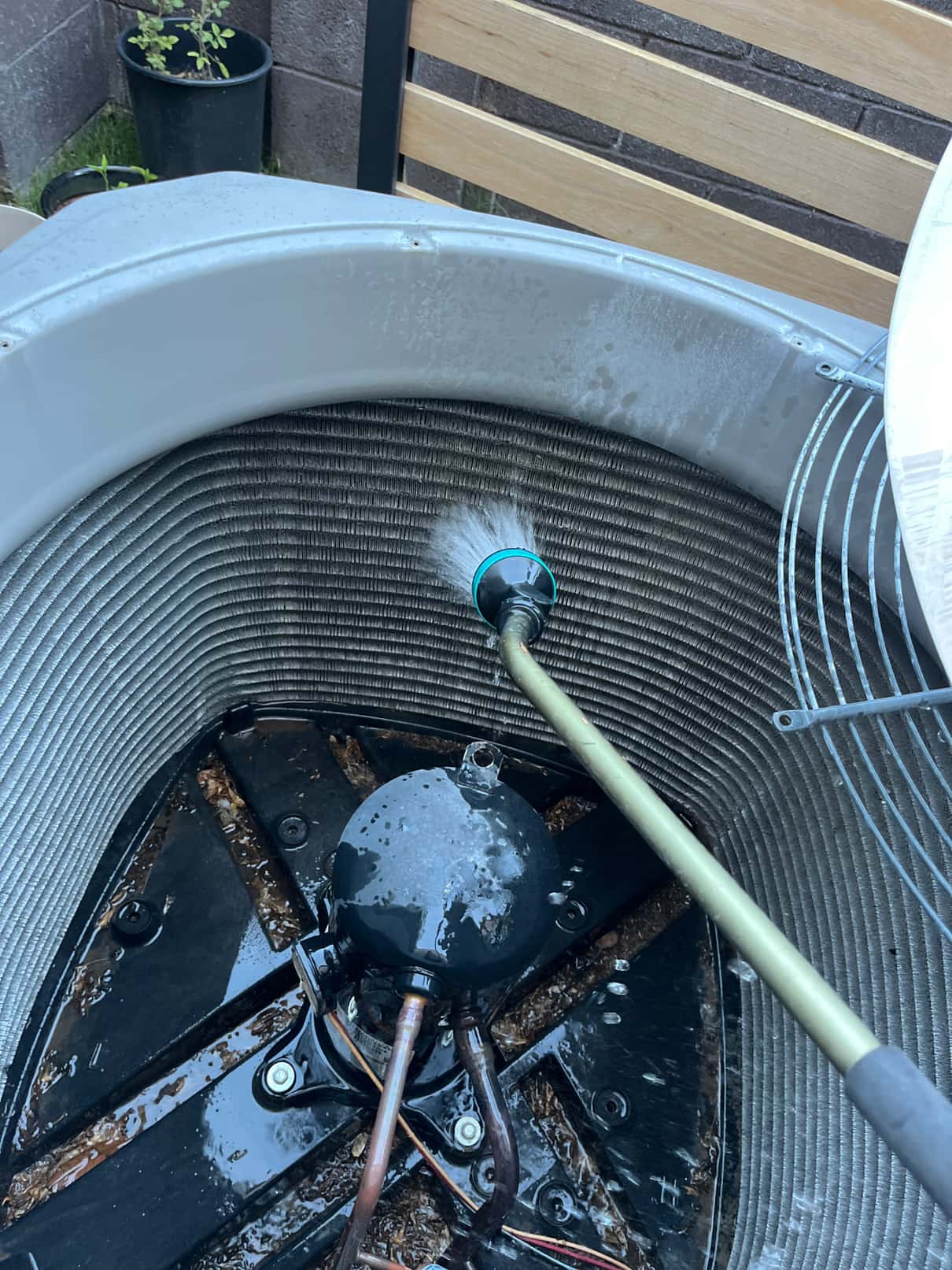 Garden hose sprays the condenser coil inside an outdoor unit, with compressor and wiring visible.
