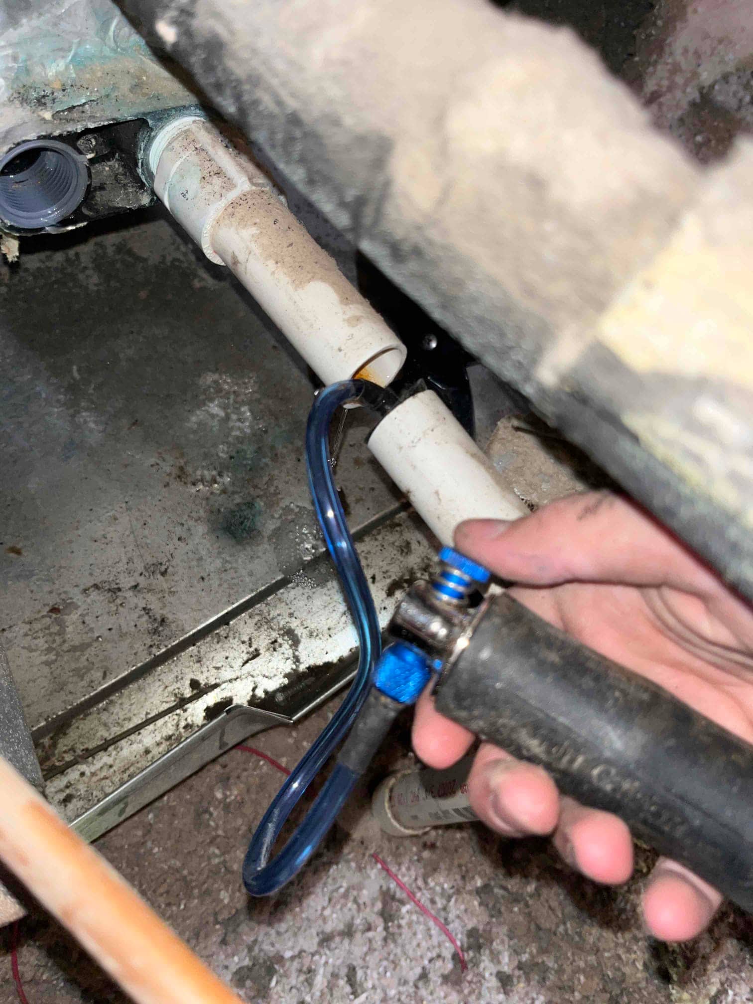 Hand holds a blow-out tool with blue hose at the condensate drain PVC connection beside the air handler’s drain pan.