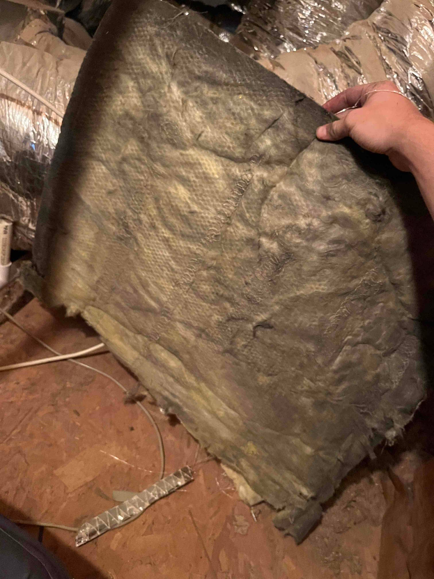 Hand holds a large, worn fiberglass panel on the attic floor beside flexible ductwork and electrical wiring.