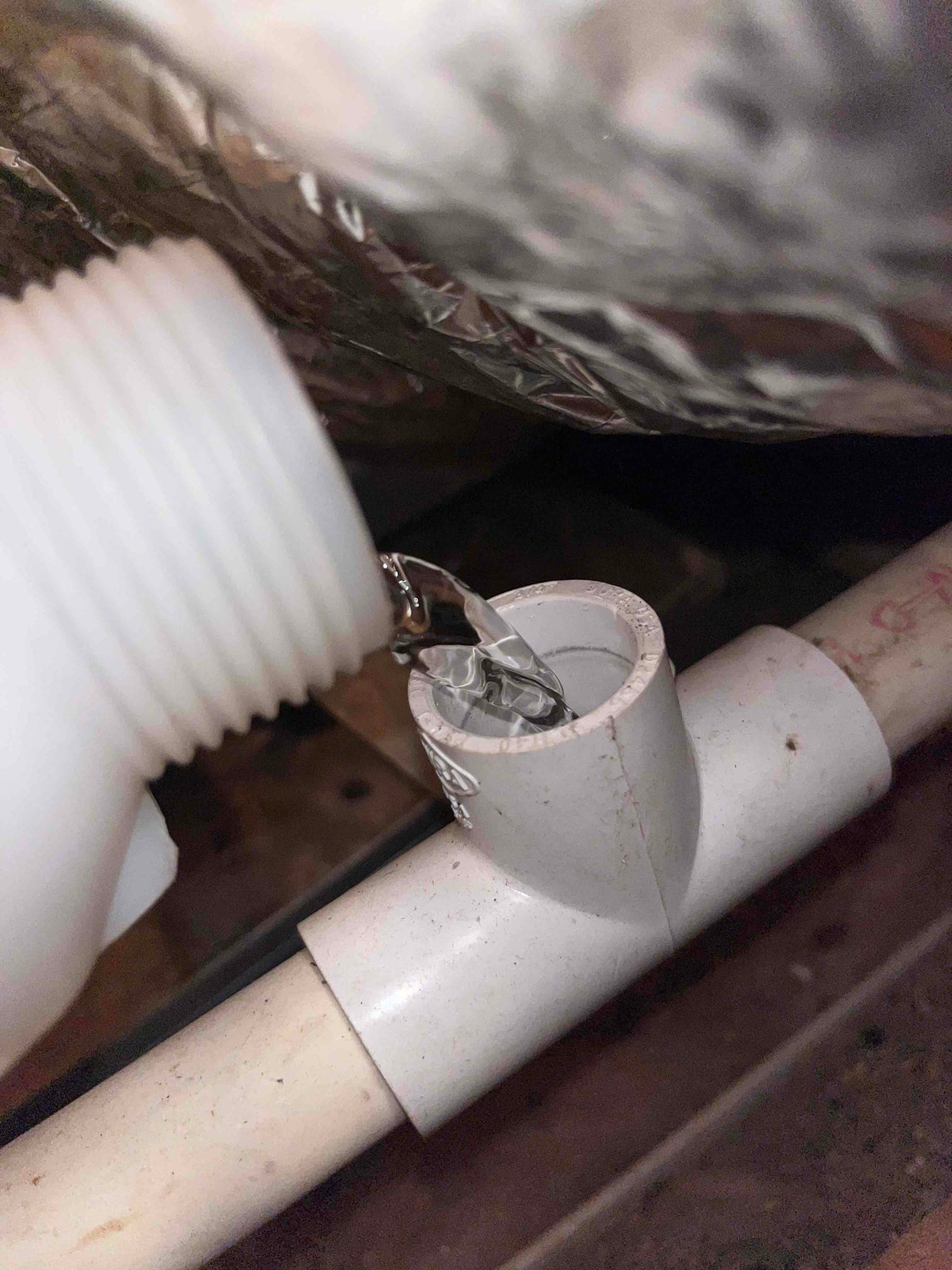 Liquid pours from a plastic container into a PVC condensate drain cleanout tee below duct insulation.