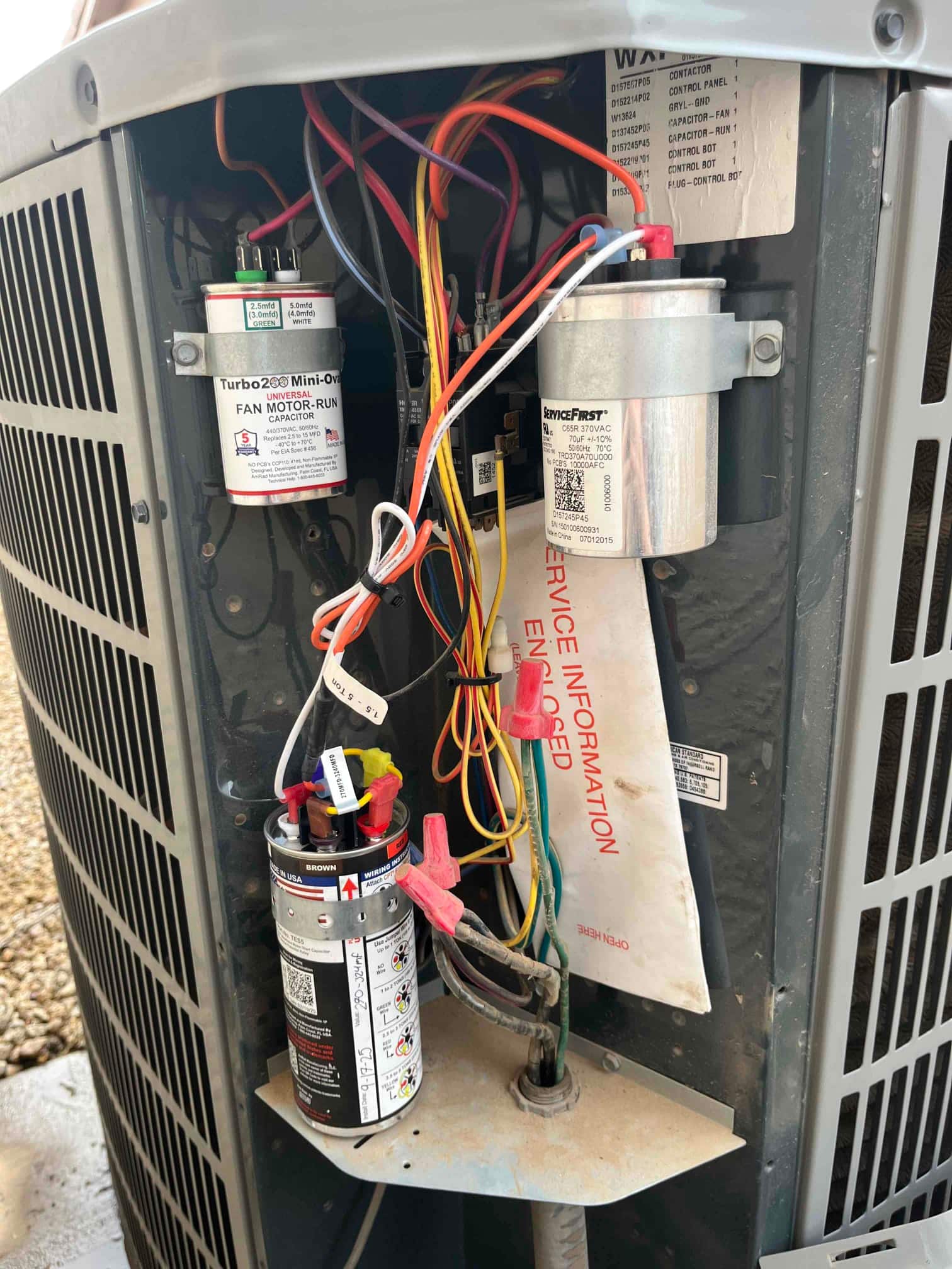 Outdoor condenser electrical compartment open, showing multiple run capacitors with connected wiring and a service information envelope.