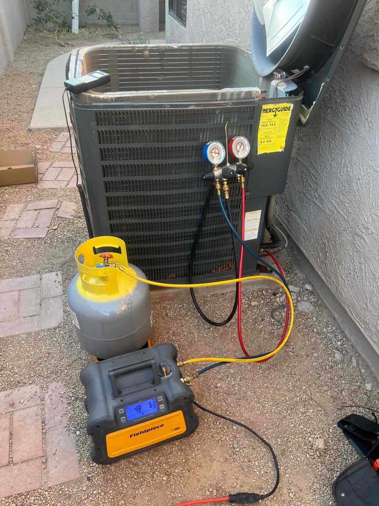 Outdoor condenser with manifold gauges connected at service ports, access panel open, and a recovery machine and refrigerant cylinder on the ground.