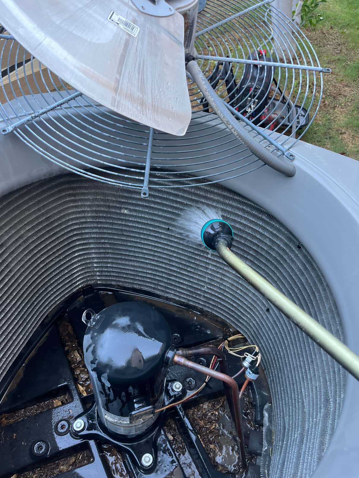 Outdoor condenser opened with the fan grille lifted while a hose rinses the condenser coil; compressor and wiring visible at the base.