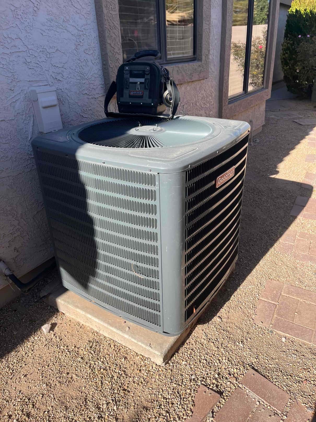 Outdoor condenser unit on a concrete pad beside the house, with a technician’s tool bag resting on top.