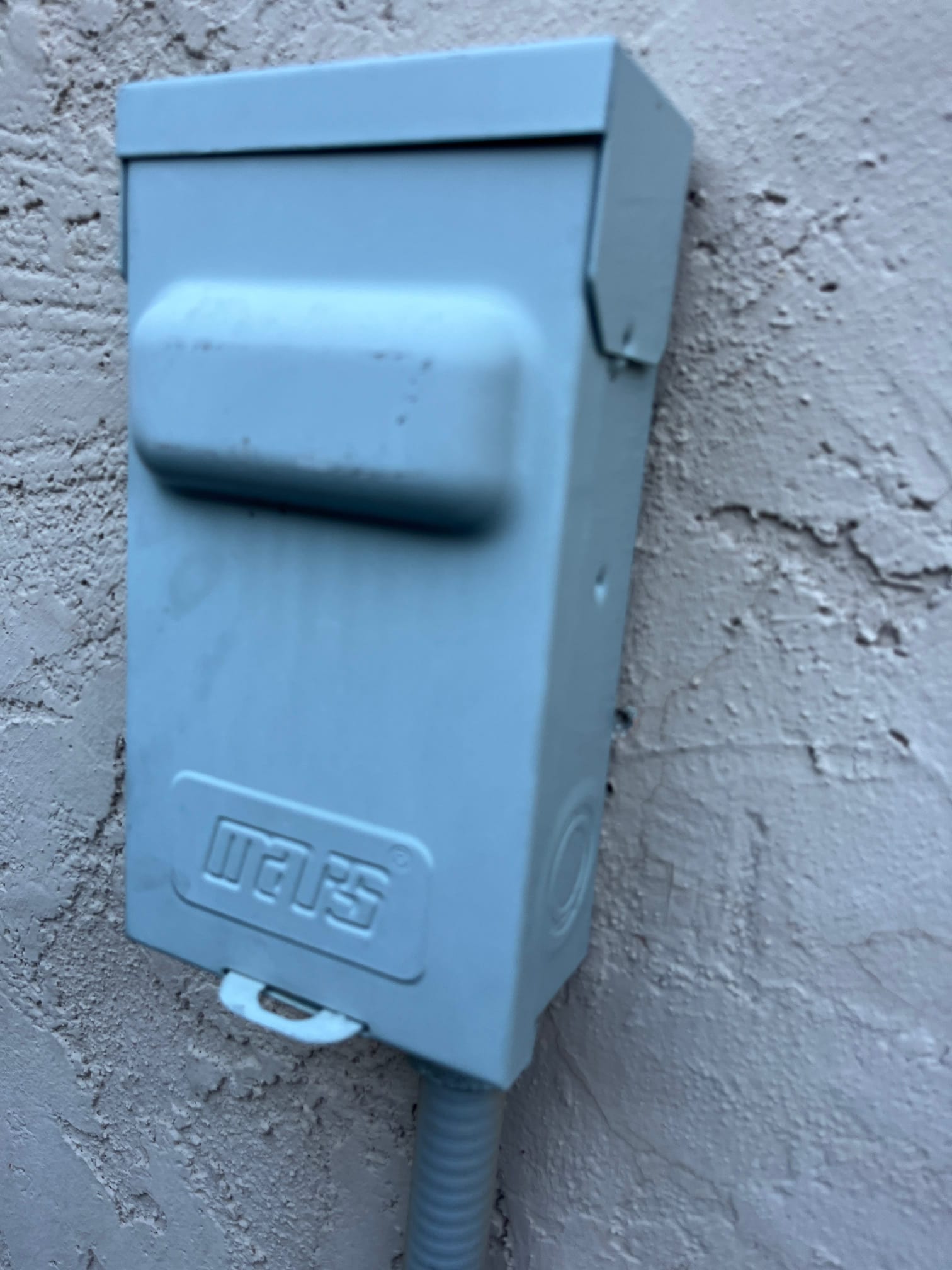 Outdoor electrical disconnect box mounted on an exterior wall with flexible conduit entering the bottom.