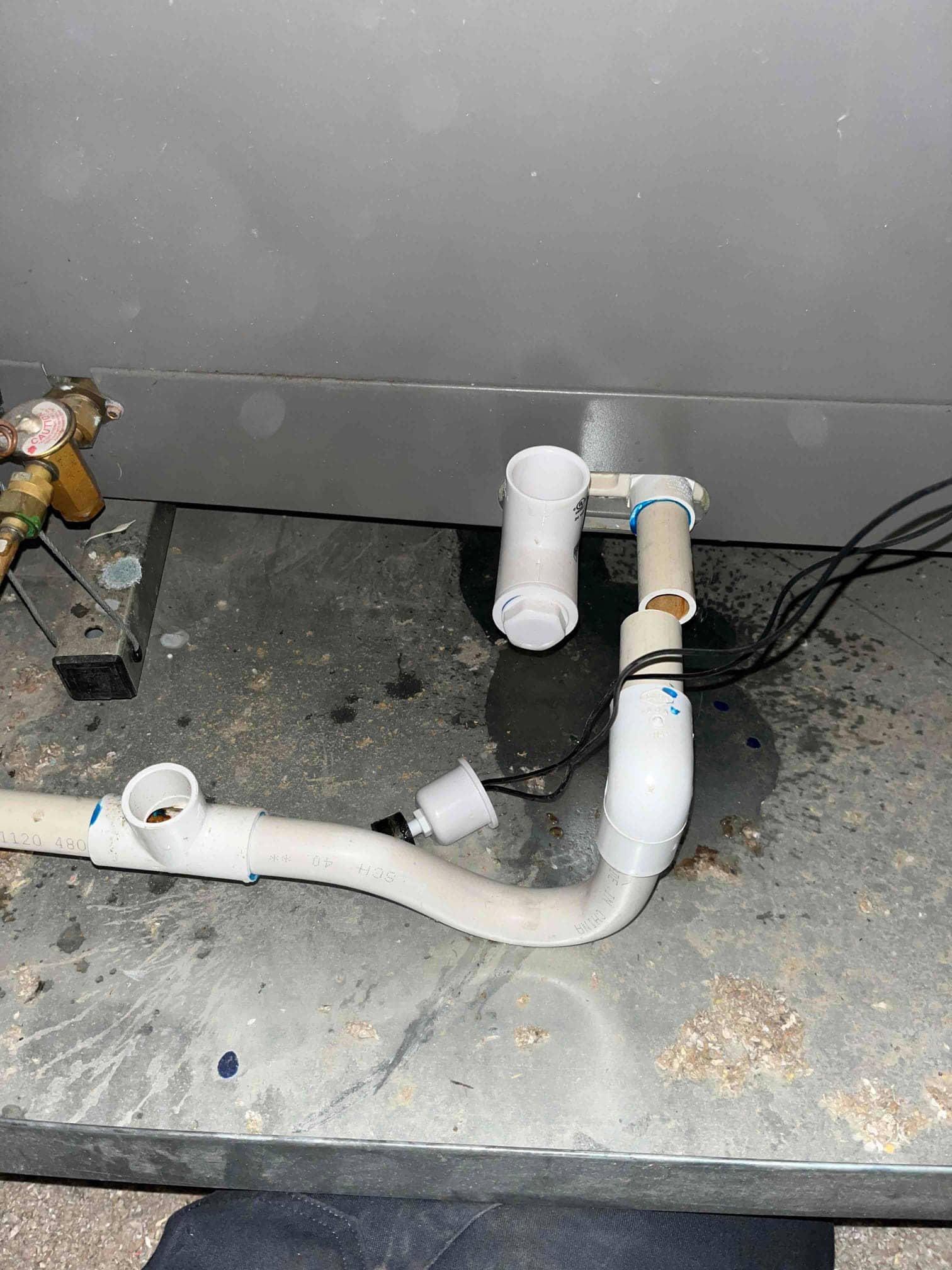 PVC condensate drain assembly with cleanout tees and inline float switch beneath an air handler, with water staining on pan surface.