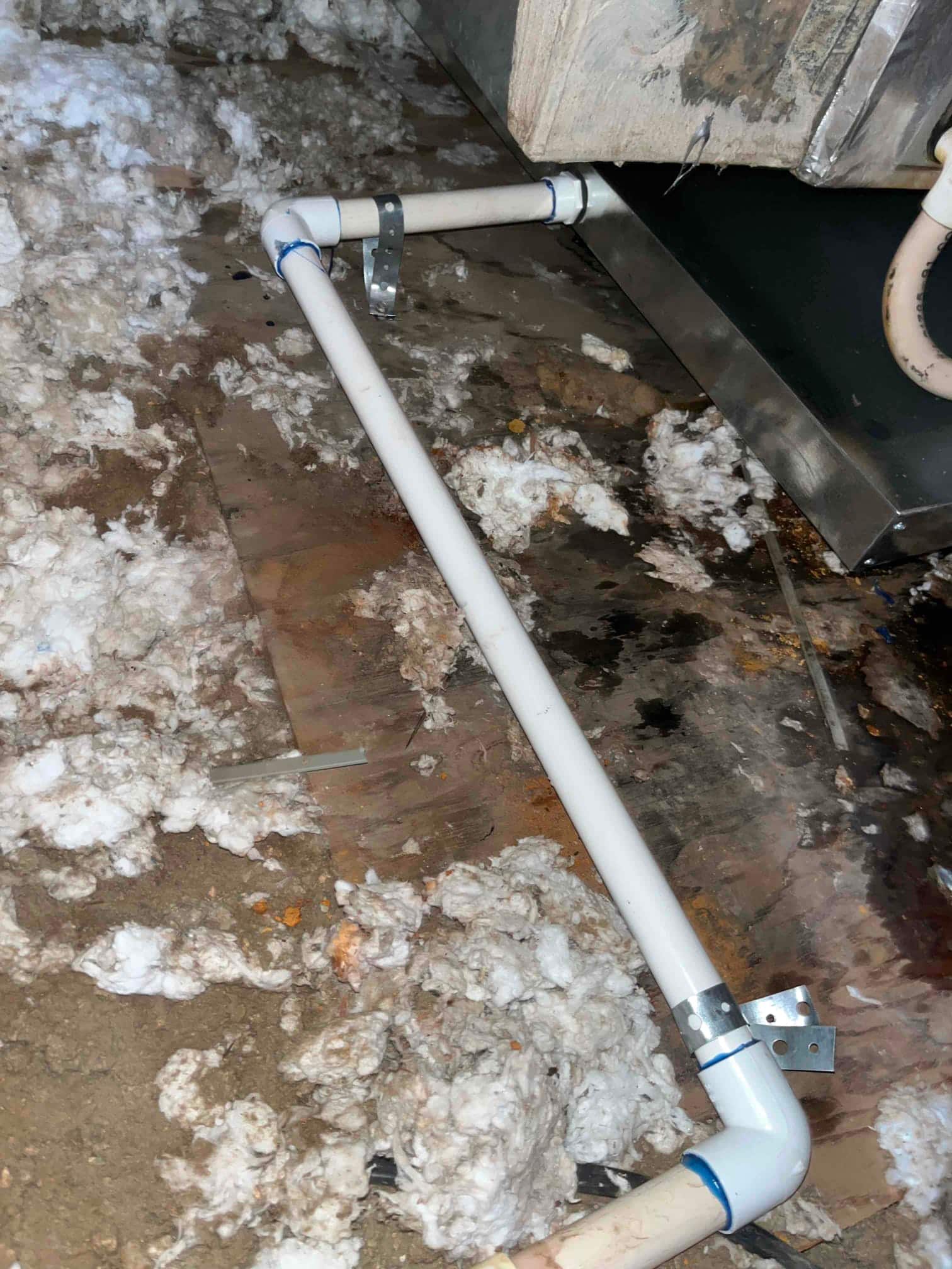 PVC condensate drain piping with elbows and metal strap supports beside an indoor unit and drain pan.