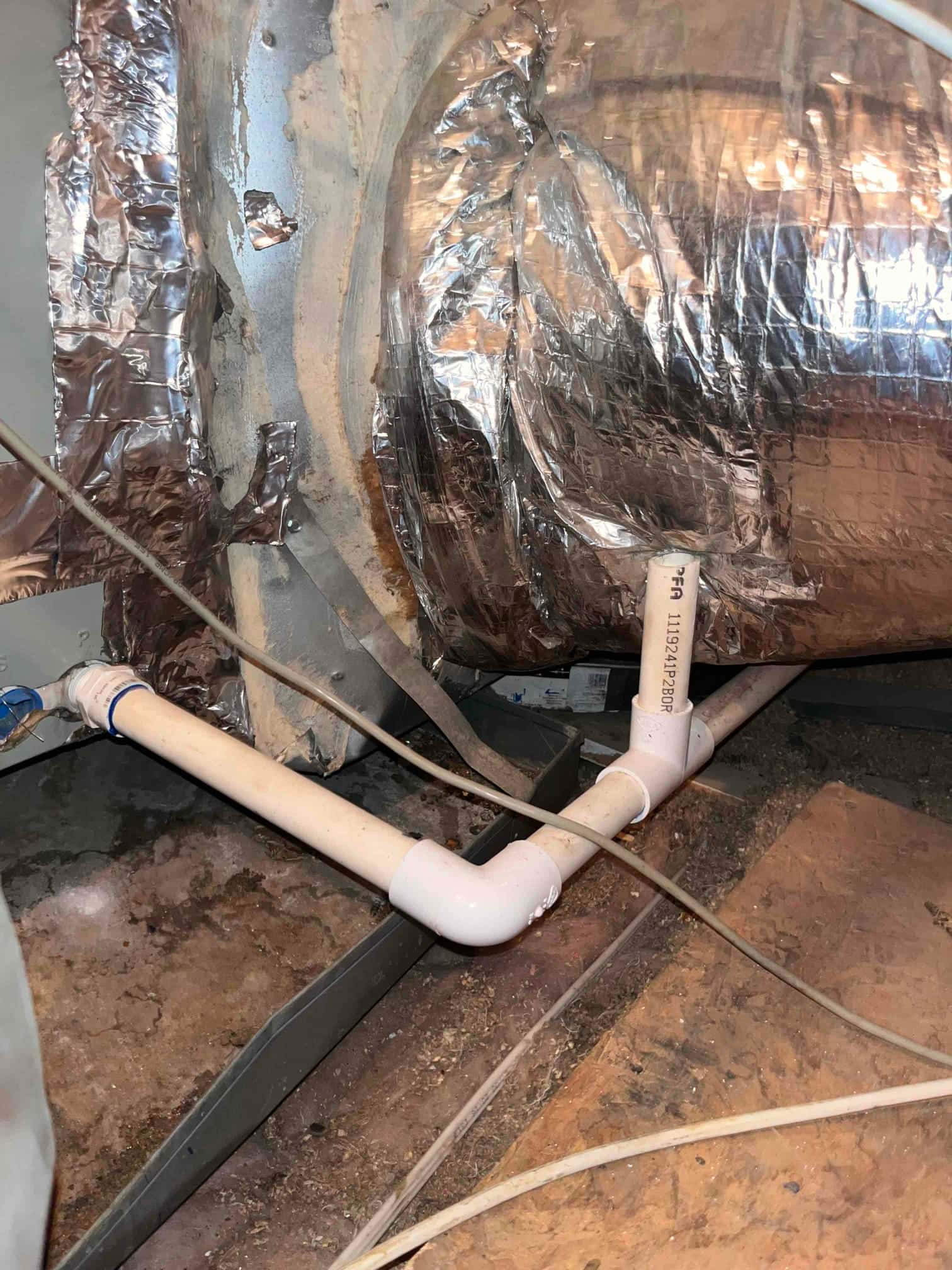 PVC condensate drain piping with tee and elbow exits an indoor air handler above an auxiliary drain pan.