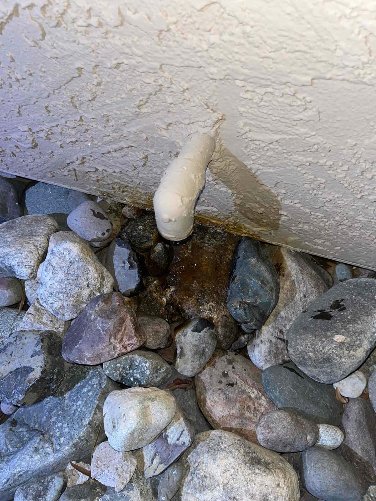 Short PVC condensate drain terminates at exterior wall, with moisture and wet staining on surrounding rocks.