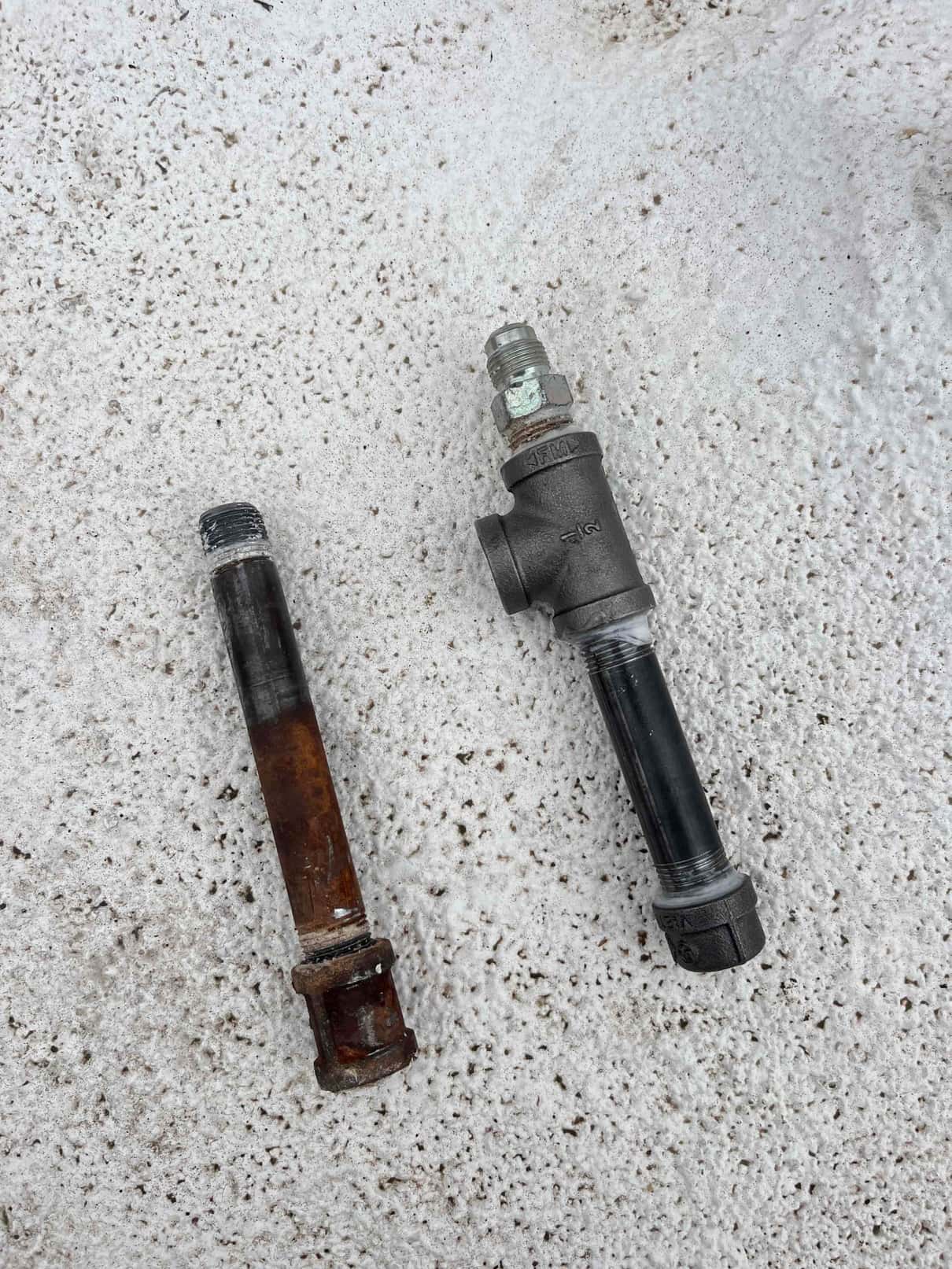 Two threaded pipe assemblies on a textured surface; one corroded and removed, another newly assembled with a tee and capped nipple.
