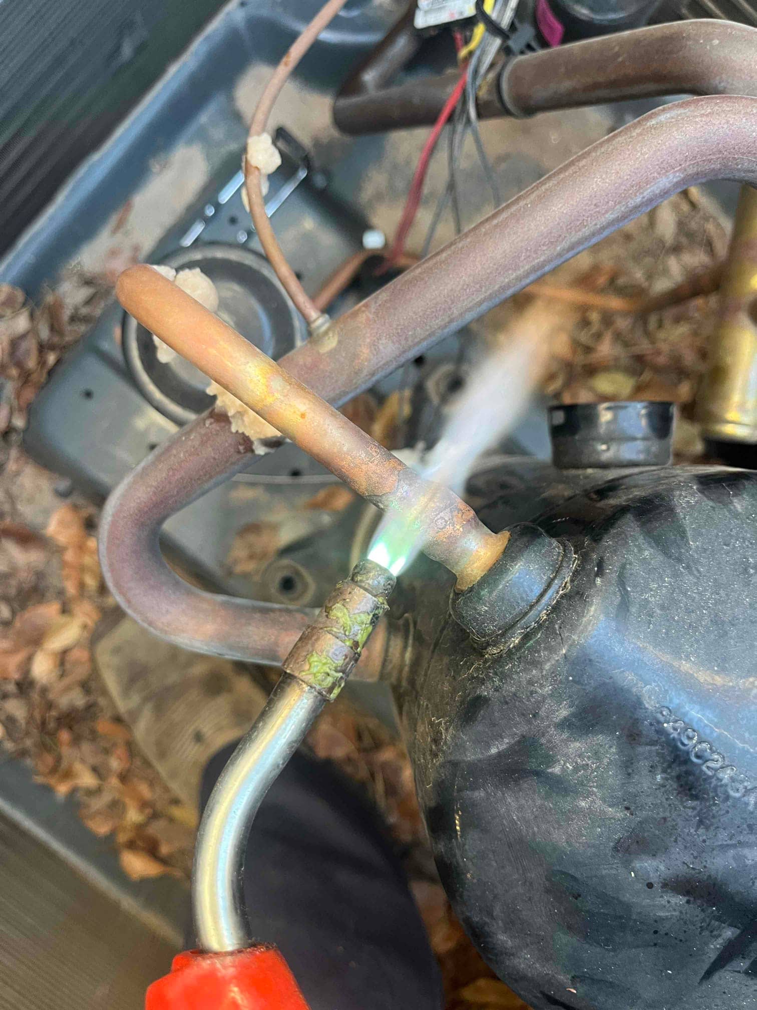 Torch heats copper refrigerant tubing at a compressor connection inside the outdoor unit, showing active brazing with visible flame.