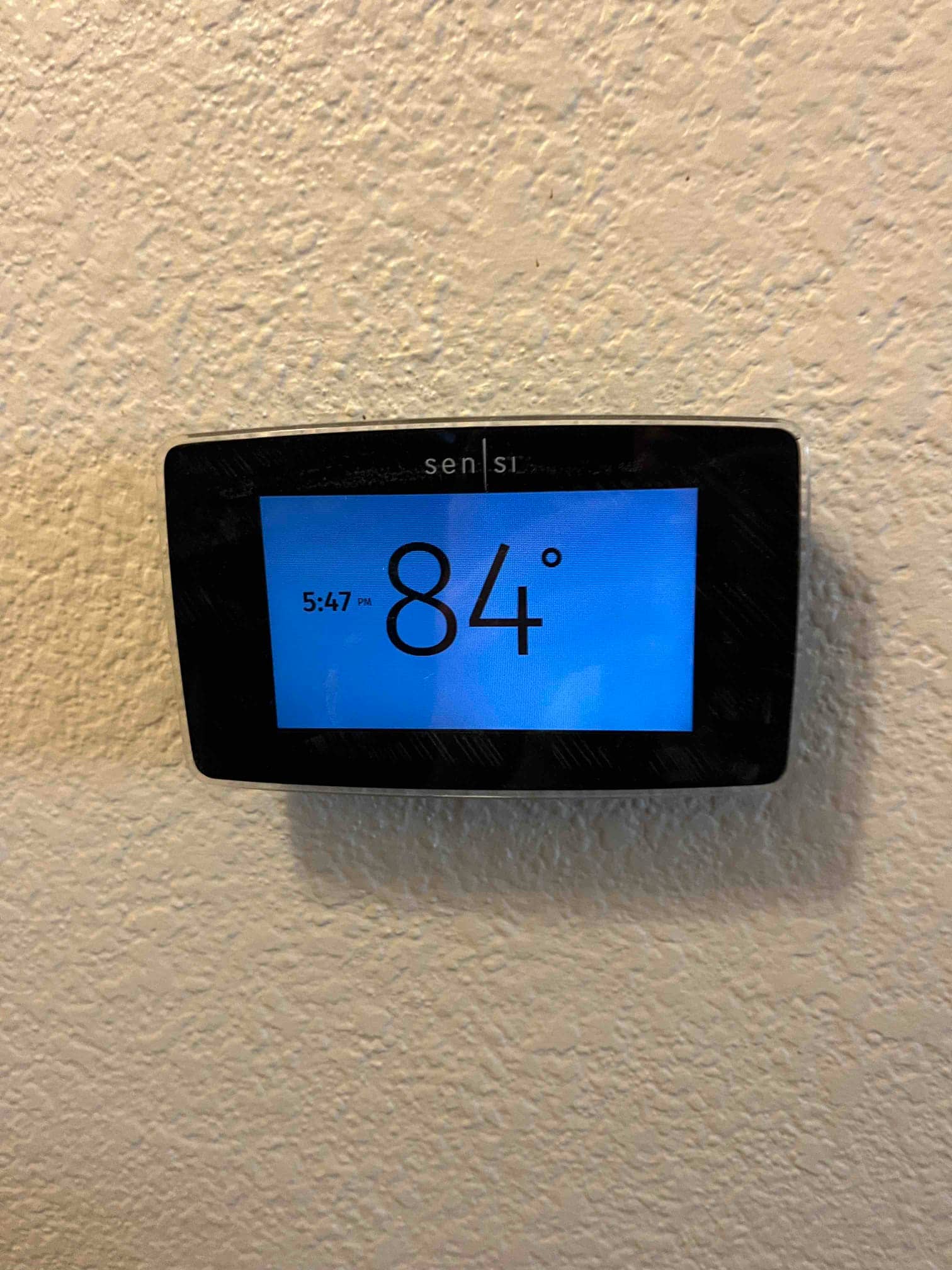 Wall-mounted digital thermostat displays 84° room temperature on blue screen with time shown at 5:47 PM.