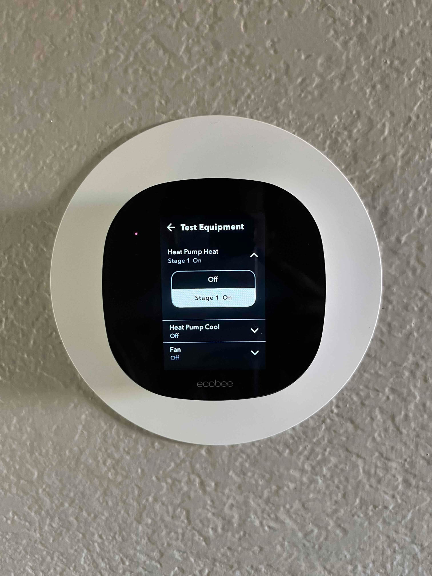 Wall-mounted smart thermostat displays Test Equipment menu with Heat Pump Heat Stage 1 On, Heat Pump Cool Off, and Fan Off.