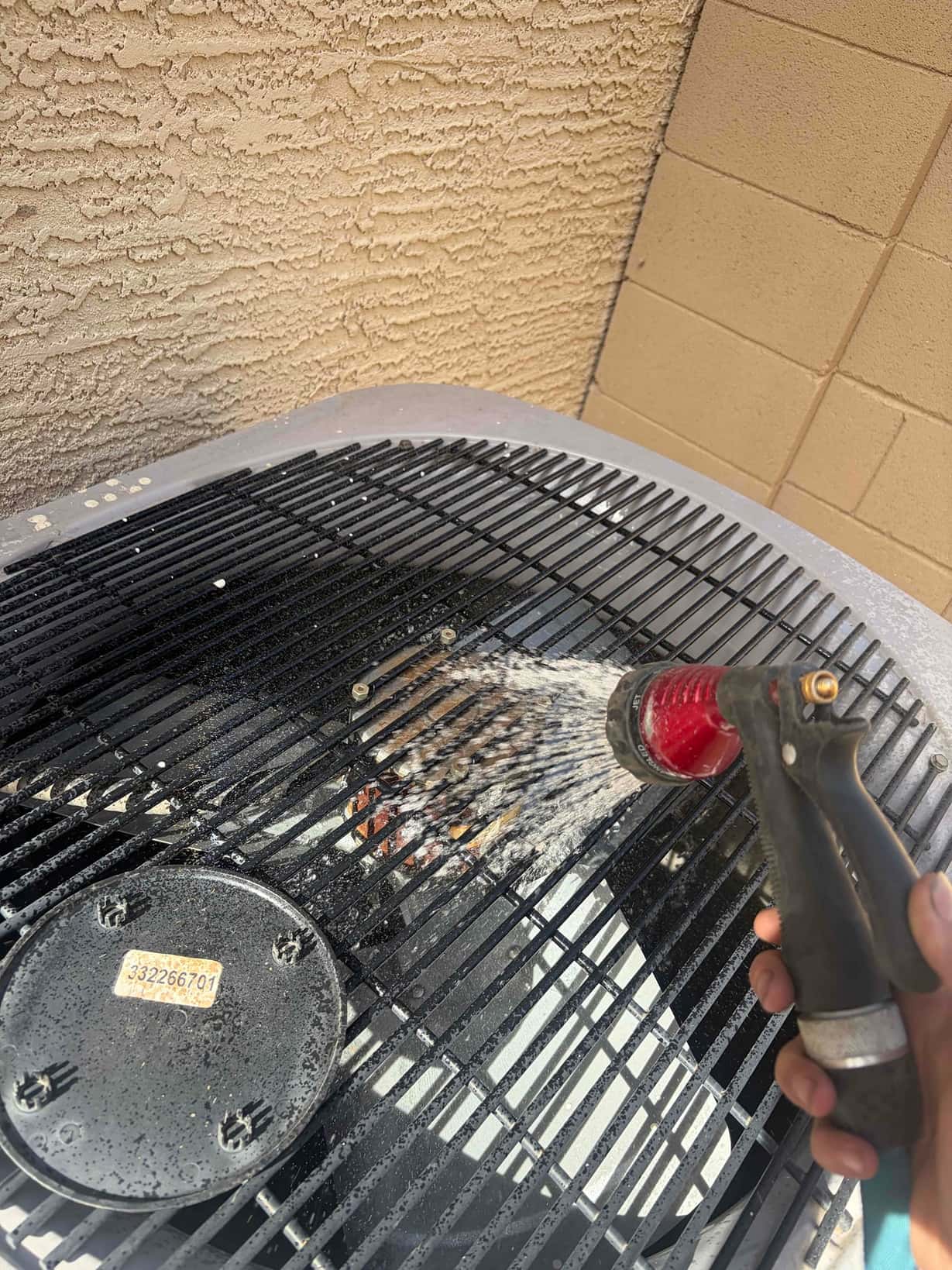 Water hose sprays through the top grille of an outdoor condenser unit, rinsing debris around the fan hub.