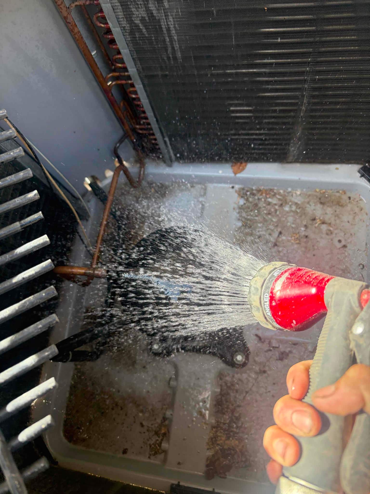 Water spray inside condenser cabinet onto base pan and coil surfaces, with debris and leaves visible.