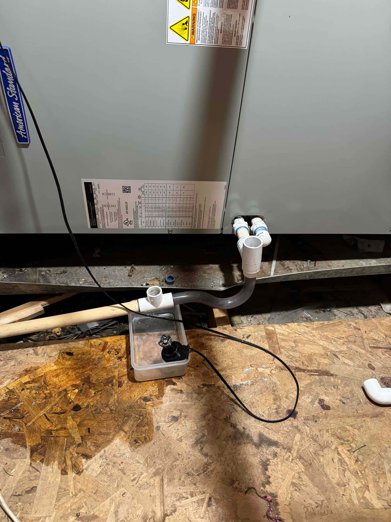 Air handler front panel with PVC condensate drain and cleanout tee, with water visible on plywood beneath the drain line.