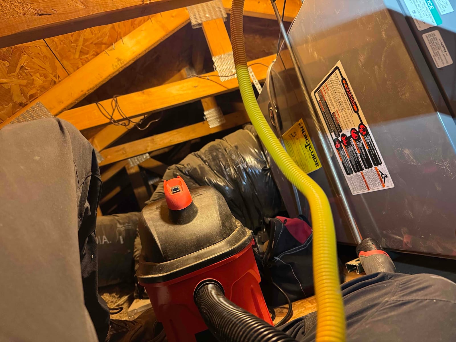 Attic installation shows a horizontal furnace or air handler with flexible ductwork, yellow gas connector, and vacuum and tools positioned nearby.