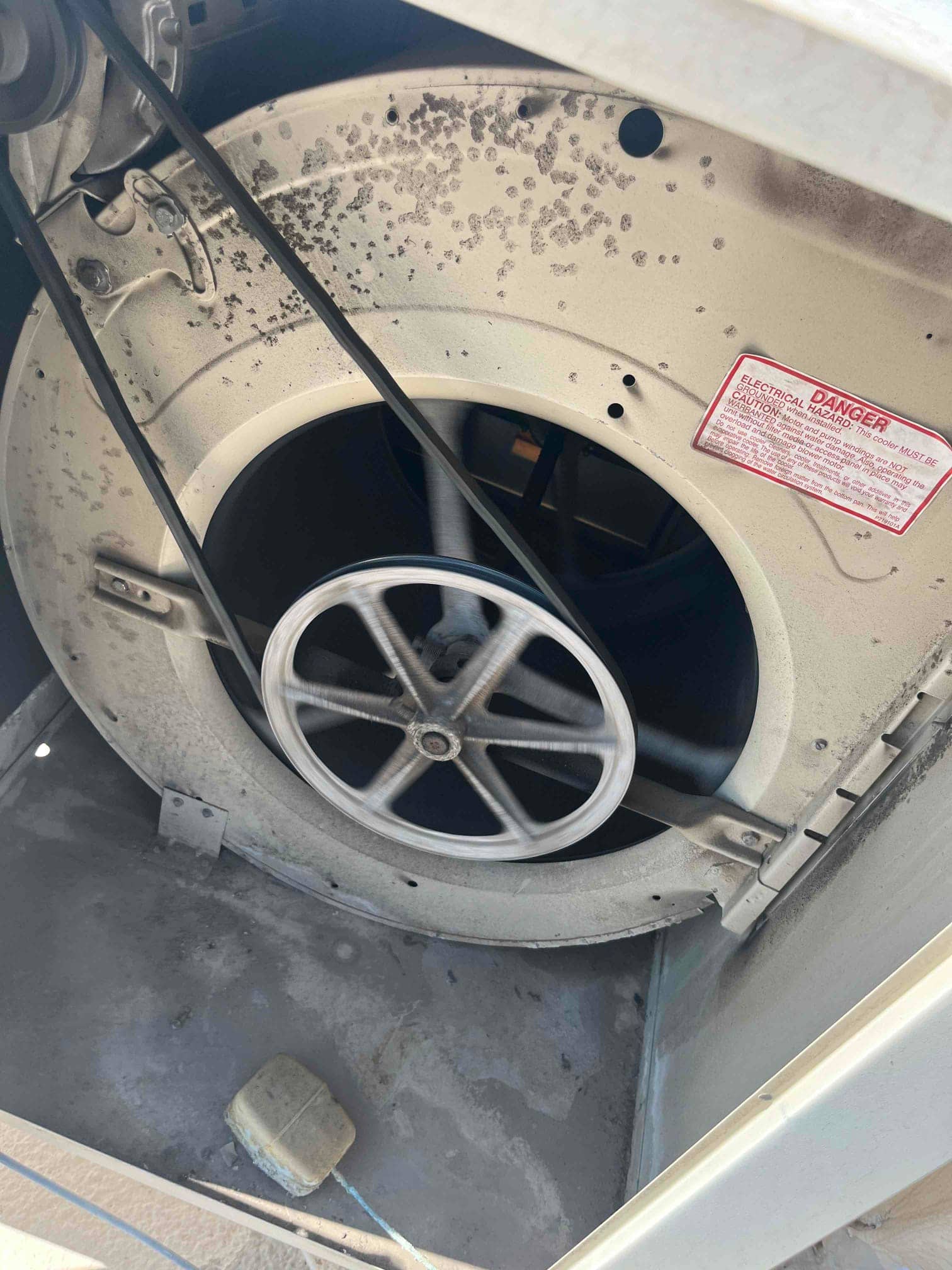 Belt-driven blower wheel and pulley rotate inside metal housing, with dust accumulation and surface corrosion visible around the shroud.