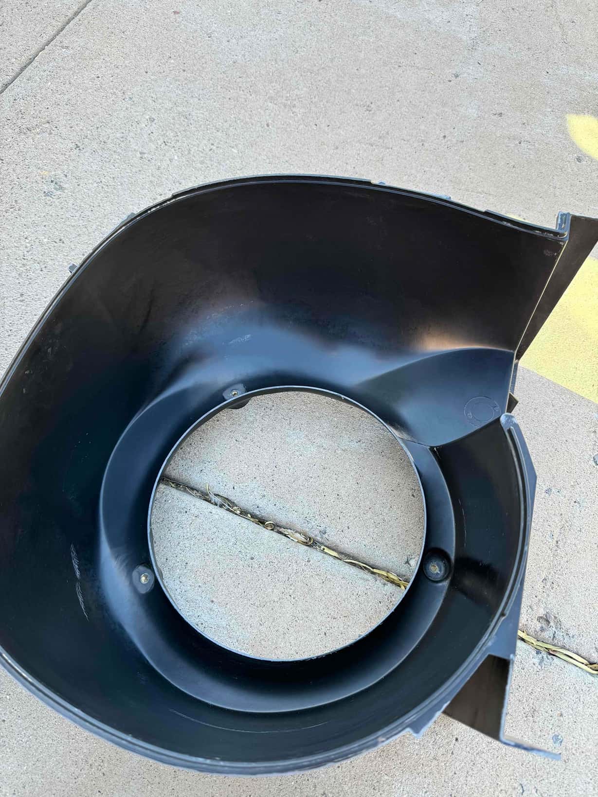 Black plastic fan shroud removed and placed on concrete, showing circular opening and molded mounting points.