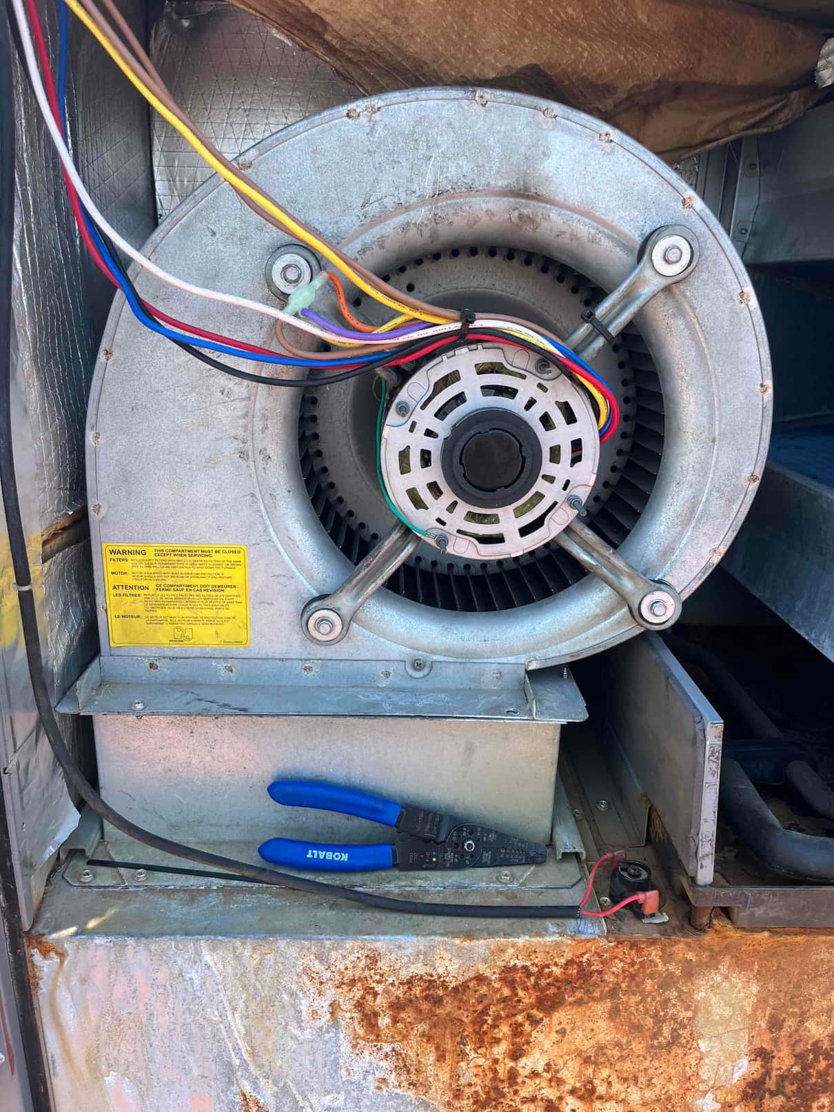 Blower motor and squirrel-cage assembly with connected wiring inside air handler, with wire strippers resting on cabinet and rusted base panel.