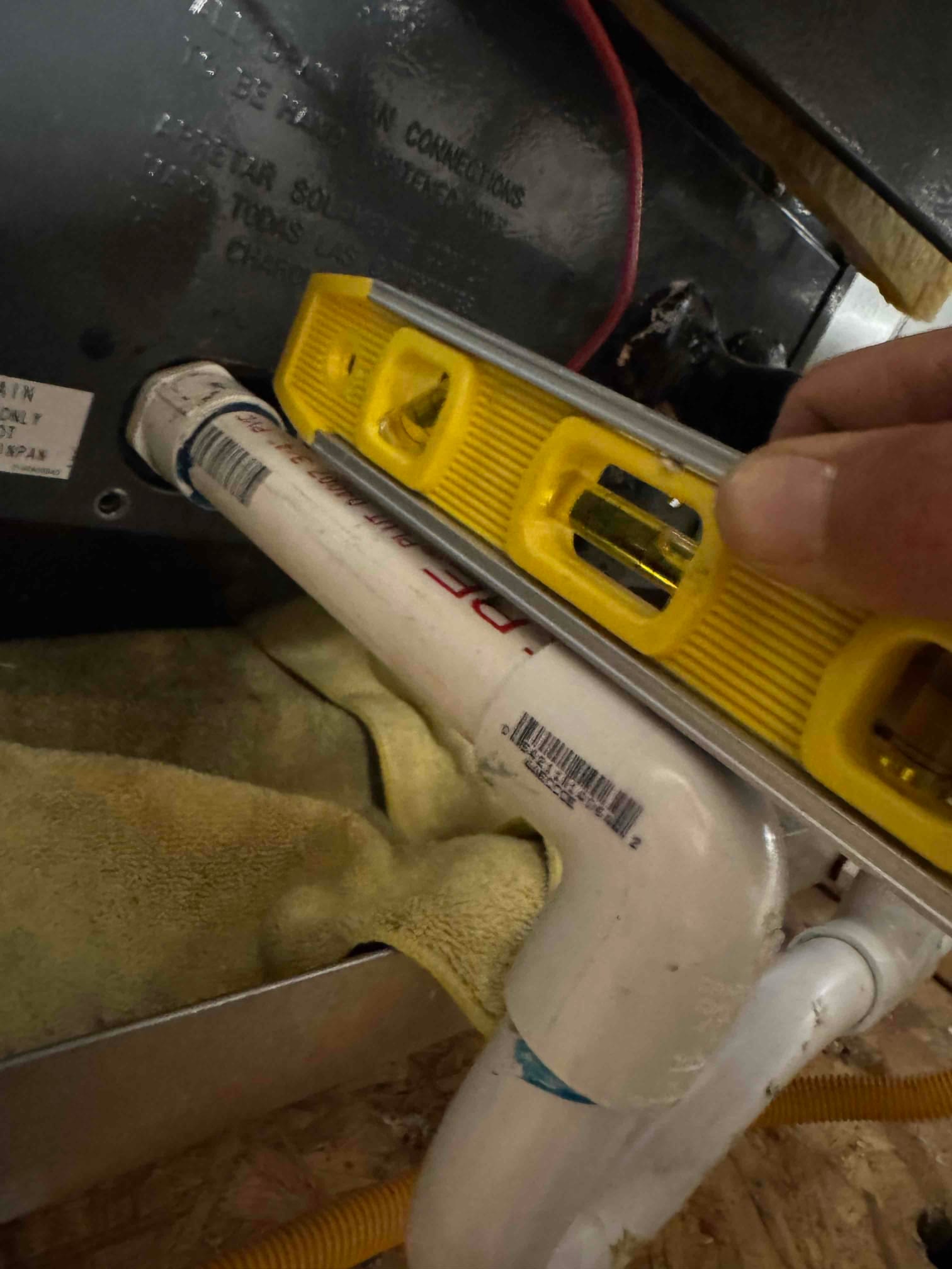 Bubble level rests on PVC condensate drain near air handler, with a hand holding it steady. Drain piping and trap visible.