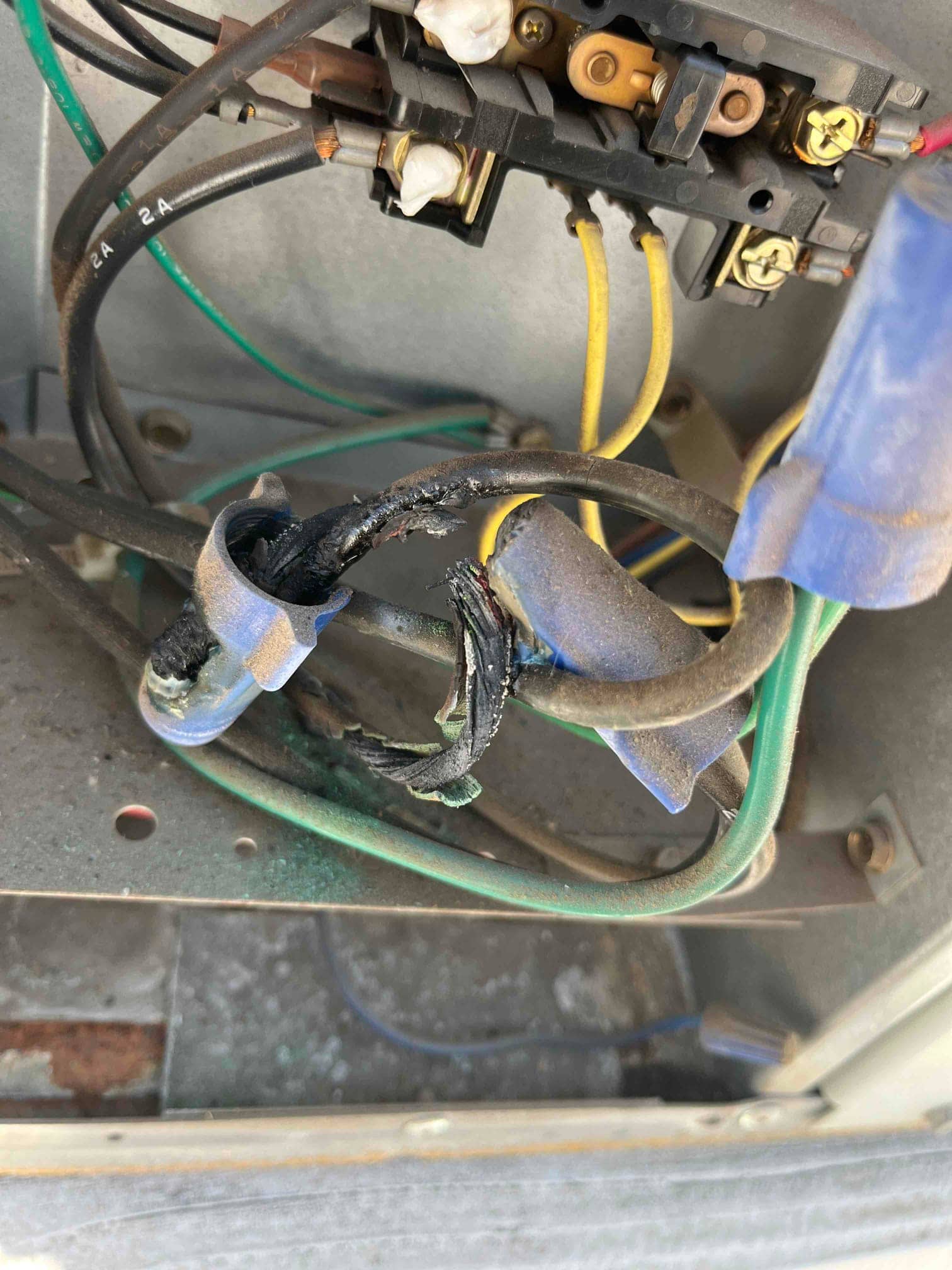 Burnt and melted wiring insulation inside outdoor condenser control compartment, below contactor, with blue connectors and green ground wire.