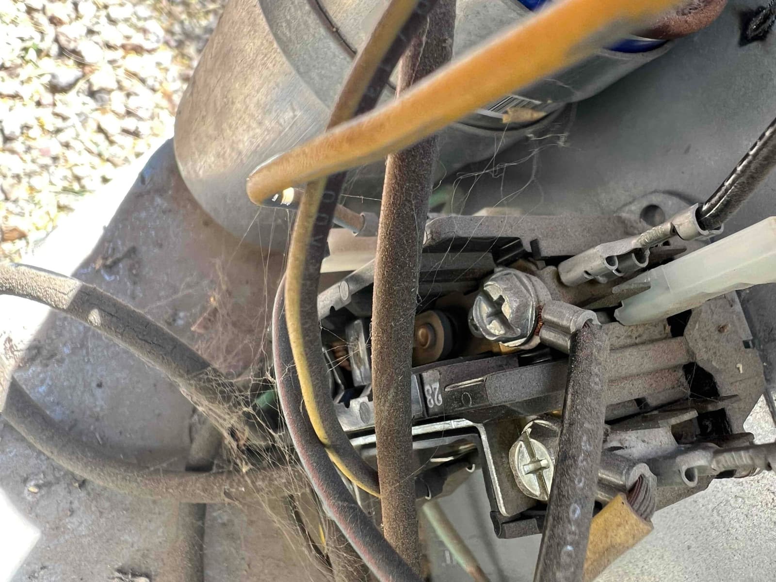 Close-up of an air conditioner contactor with connected wires and terminal screws, showing dust and cobwebs; capacitor visible in background.