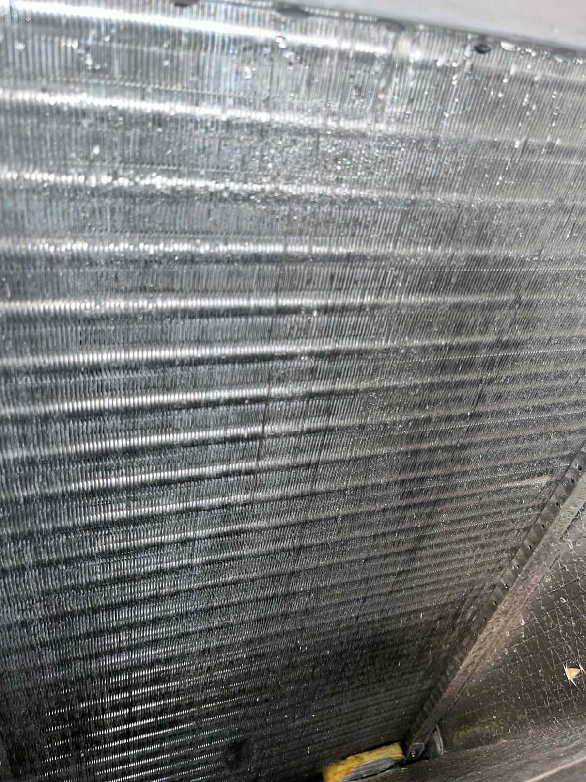Close-up of an indoor evaporator coil, showing wet aluminum fins with condensation droplets across the A-coil surface.