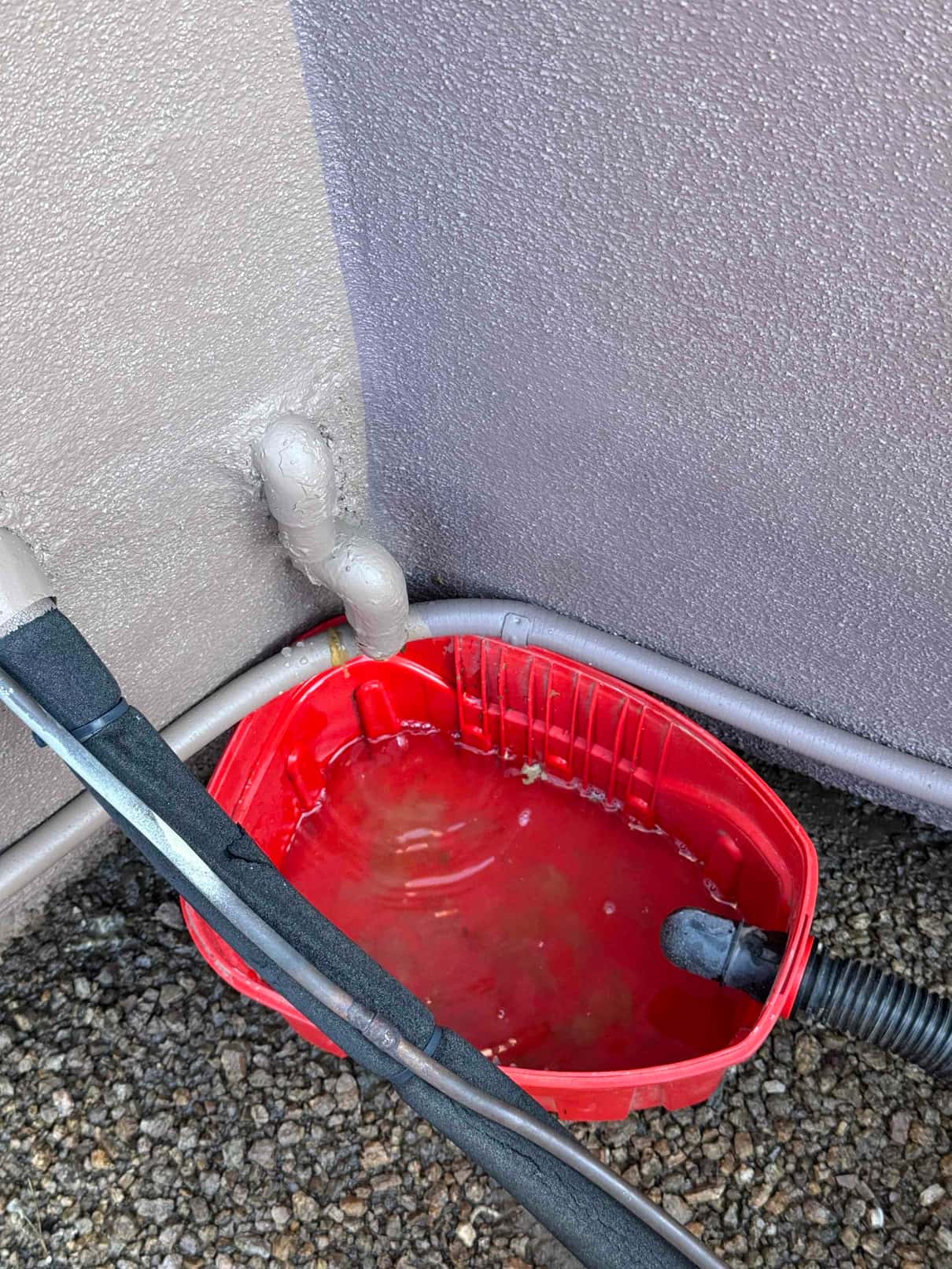 Condensate drain line discharges water into a red catch pan beside insulated refrigerant lines outside the unit.
