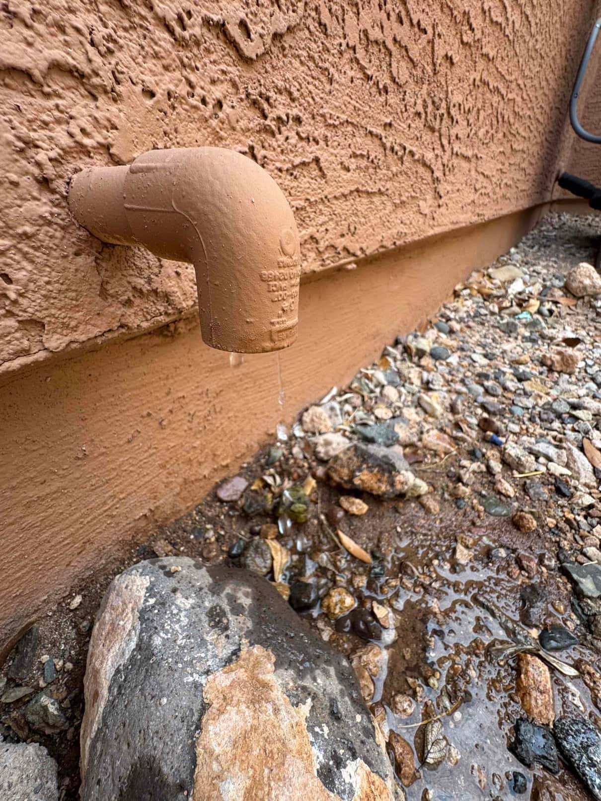 Condensate drain line terminates outside a stucco wall, with water dripping onto rocks below.