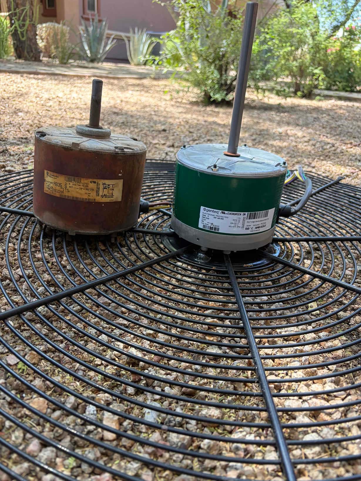 Two condenser fan motors rest on an outdoor unit fan guard, one heavily rusted and one new green motor.