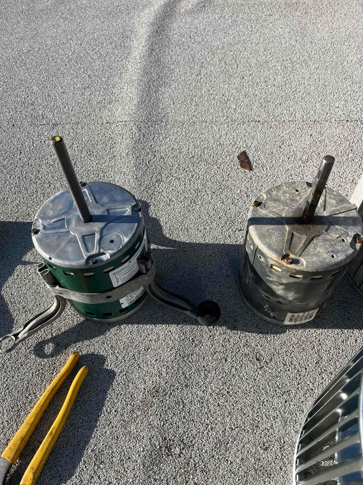 Two condenser fan motors on a rooftop surface, one with visible rust, alongside a fan guard and yellow-handled pliers.