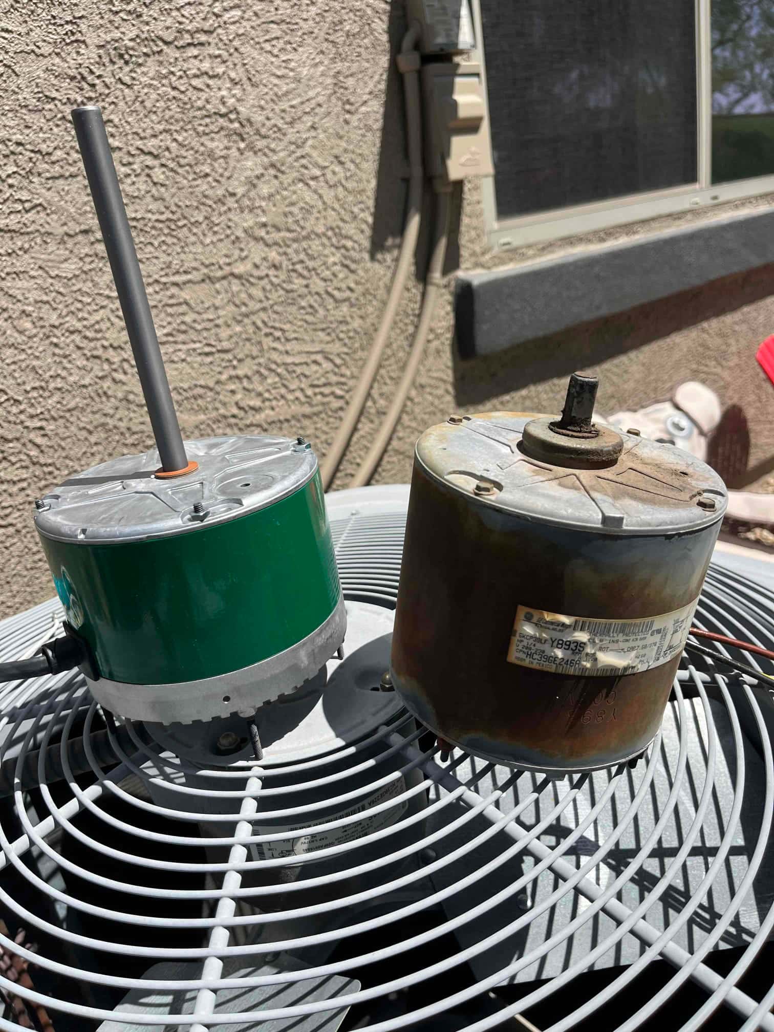 Two condenser fan motors sit on the outdoor condenser grille, one clean with long shaft, one rusted with short shaft.
