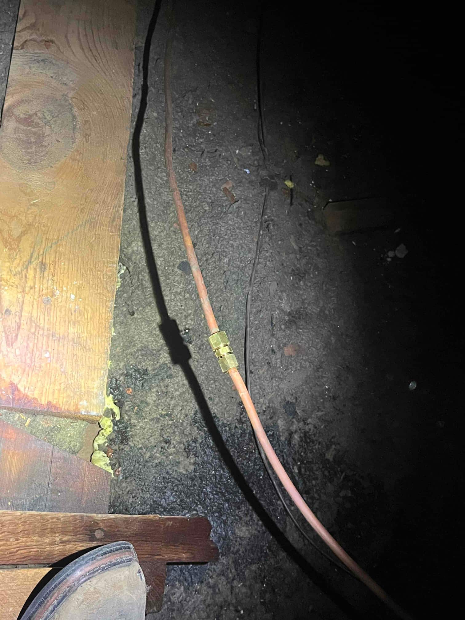 Copper tubing with a brass inline coupling runs across the crawlspace floor beside wood framing.