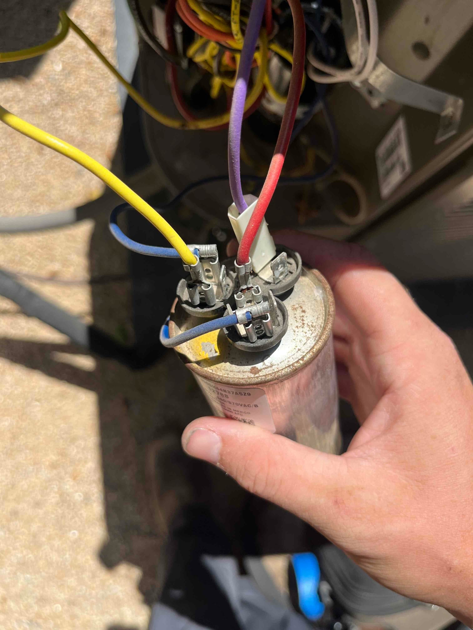 Dual-run capacitor held at an outdoor unit, showing connected purple, red, yellow, and blue wires on terminals.