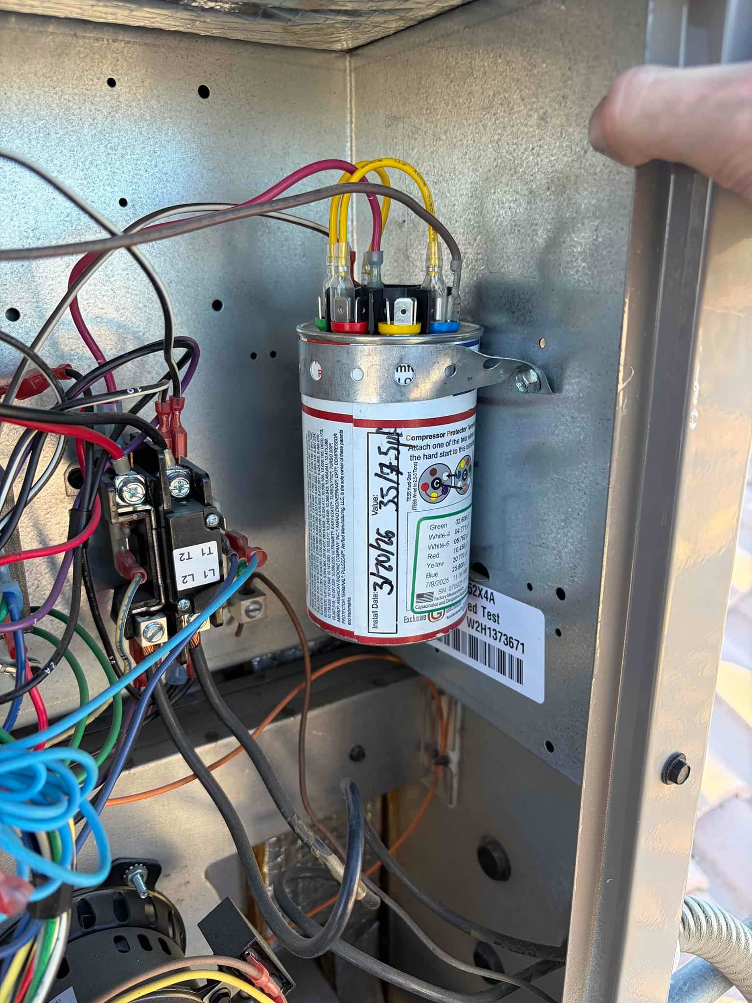 Dual-run capacitor mounted inside the outdoor unit control compartment, with yellow, red, and brown wires attached to terminals.