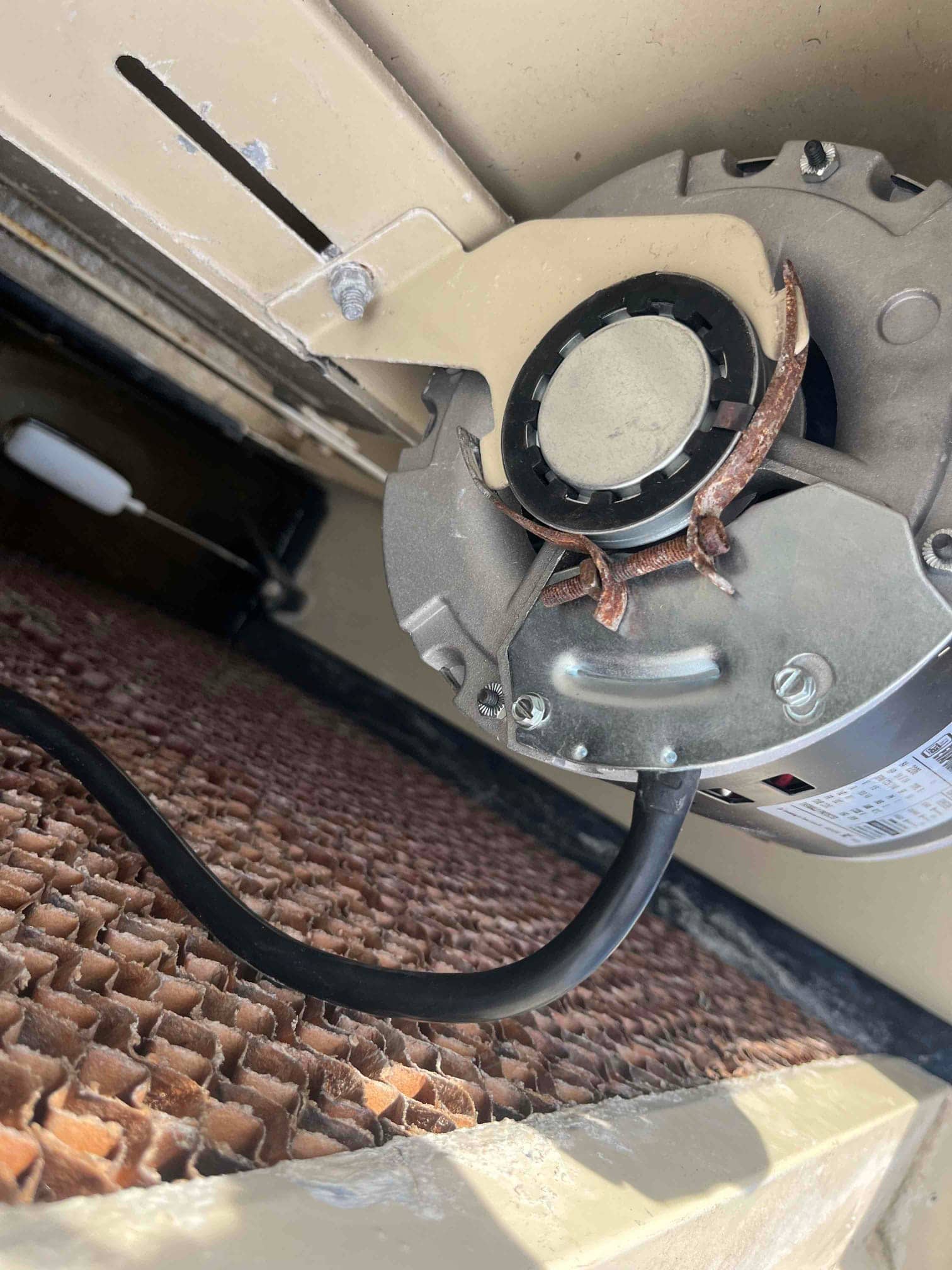 Electric motor assembly mounted inside HVAC unit above evaporative media, showing rusted clamp hardware and attached black power cable.