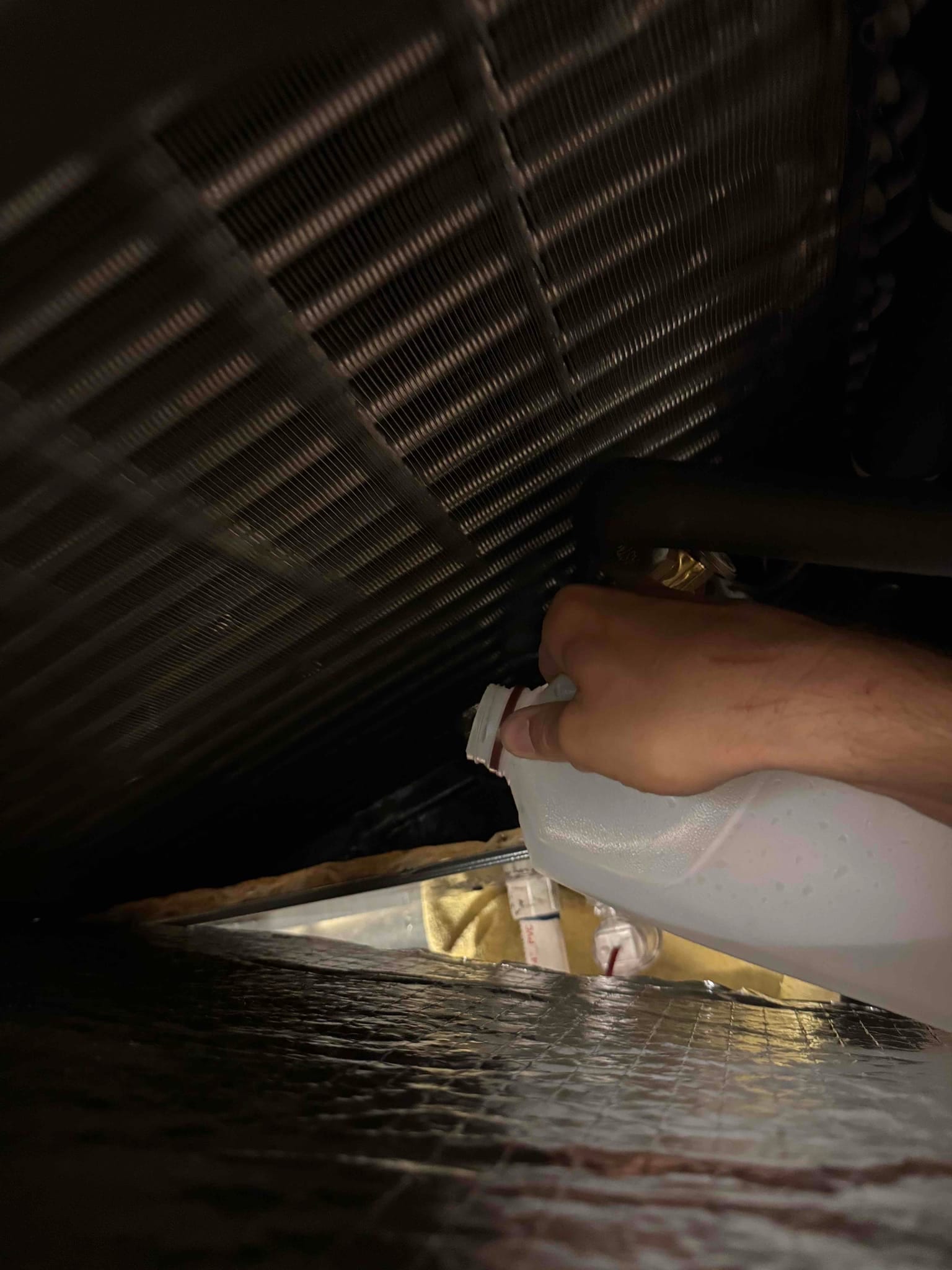 Evaporator coil fins are visible while a hand holds a plastic jug beneath the coil inside the air handler.