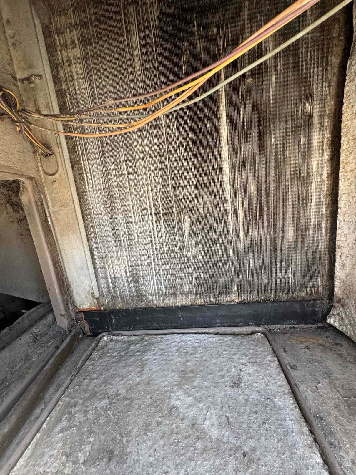 Evaporator coil fins inside an air handler show dark surface discoloration, with wiring along the side and the condensate pan below.