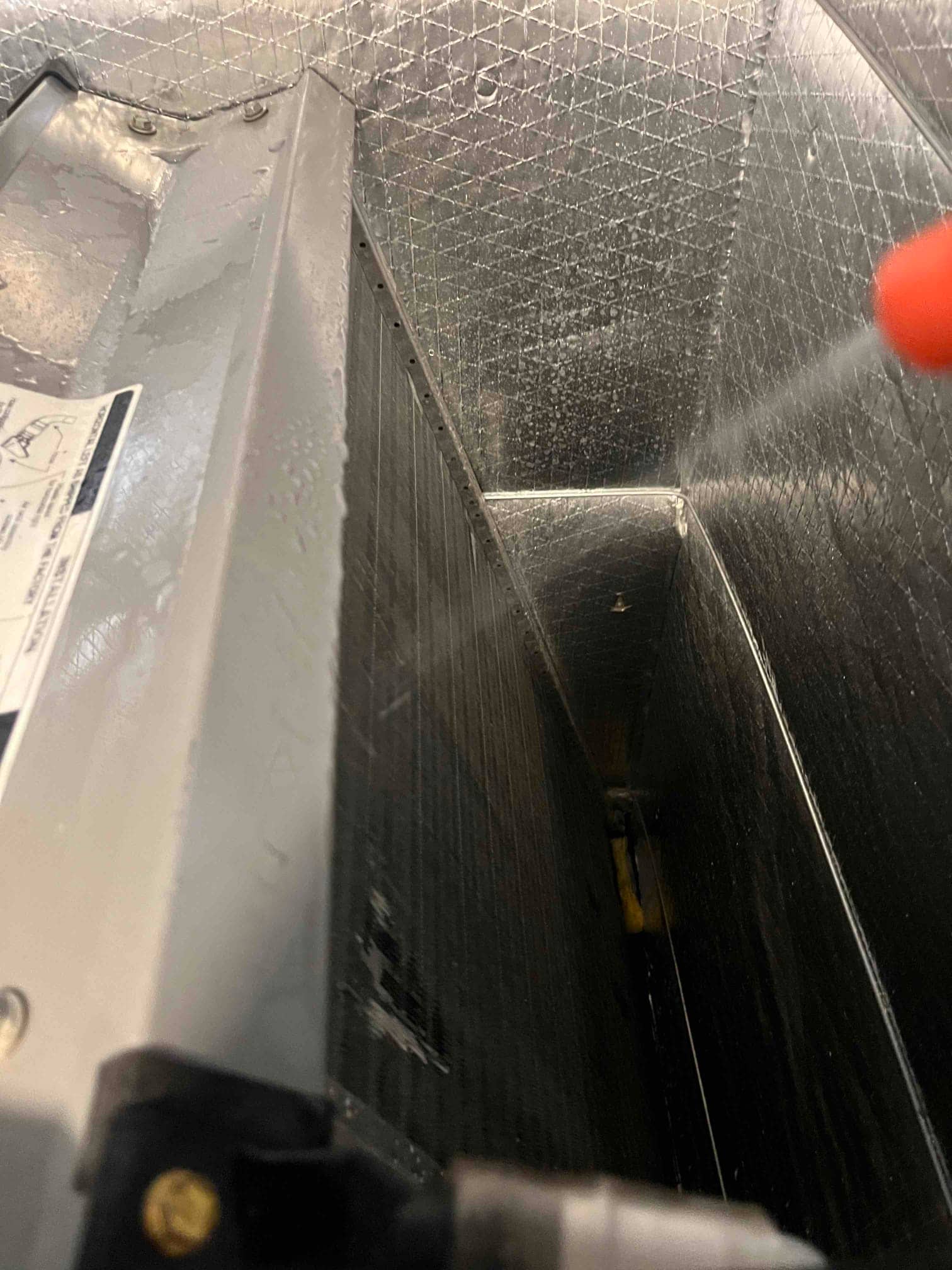 Evaporator A-coil inside air handler being rinsed with spray, with wet interior duct liner and metal housing visible.