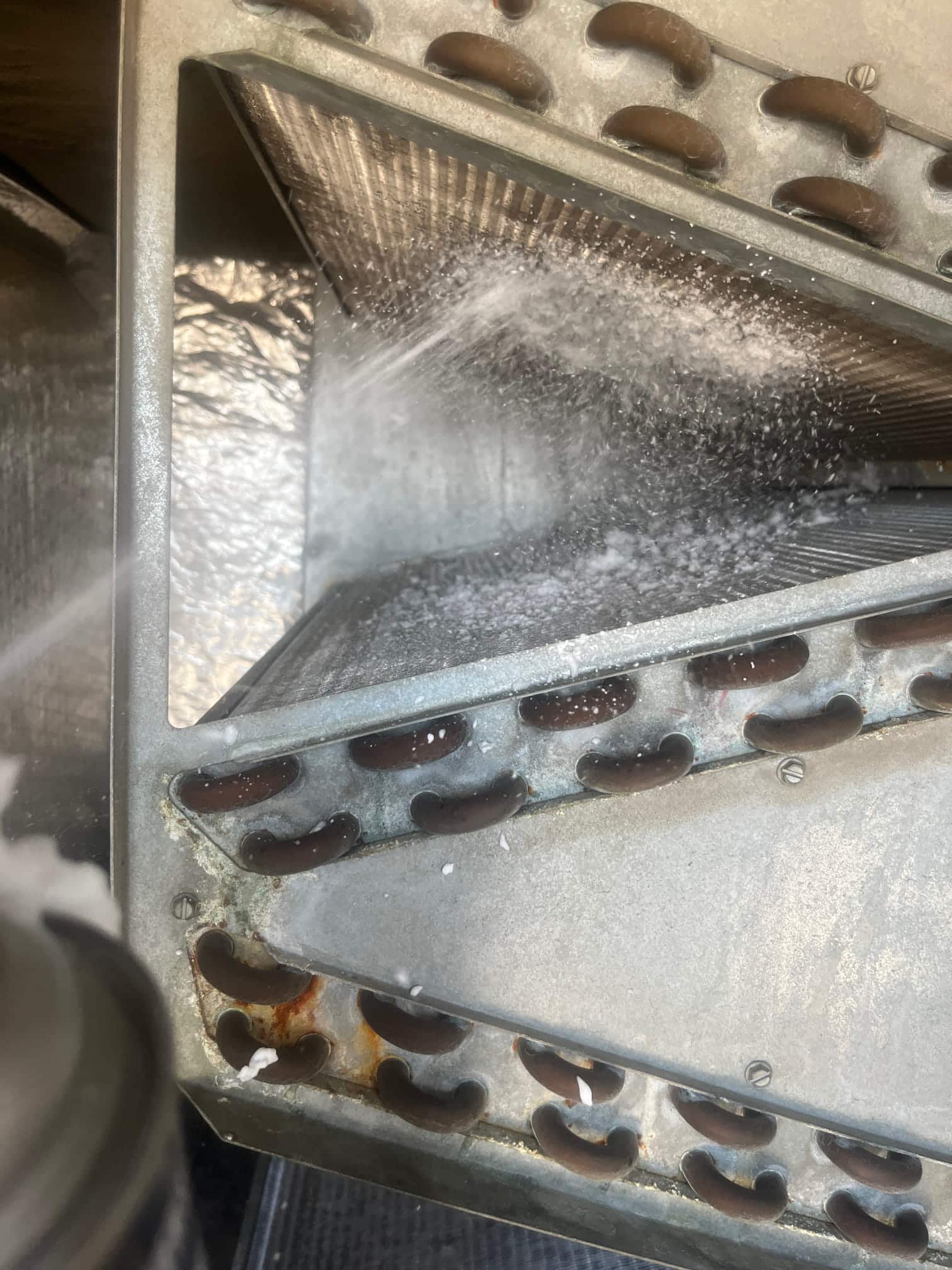 Evaporator coil section with foaming cleaner sprayed across fins and copper U-bends, light rust visible.