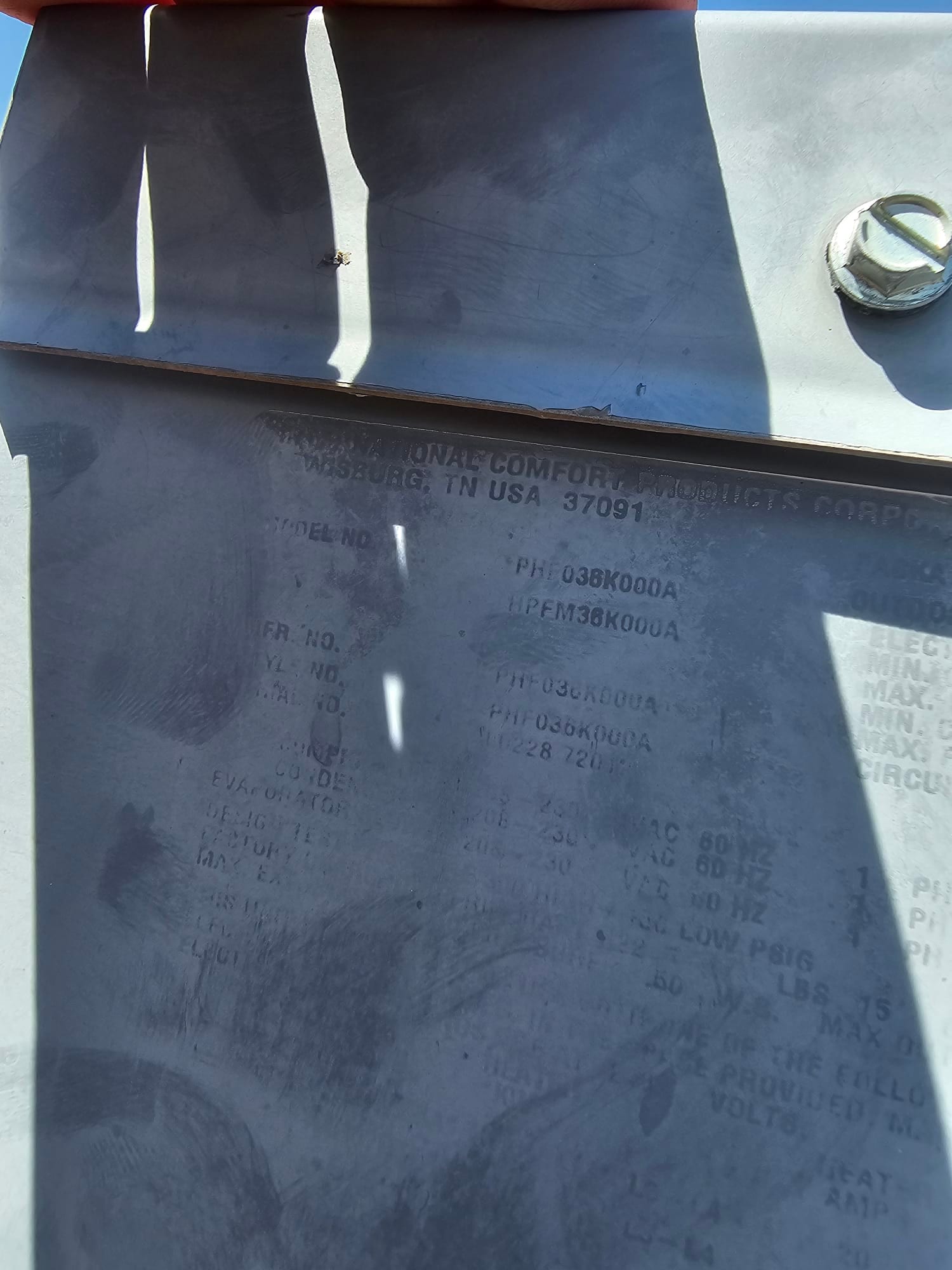 Faded equipment data plate on outdoor condenser housing with heavy smudges and shadow across the panel.
