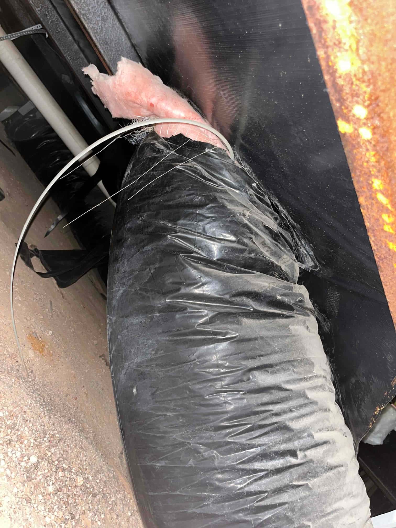 Flexible duct at an equipment connection with pink insulation exposed and a loose metal strap hanging.