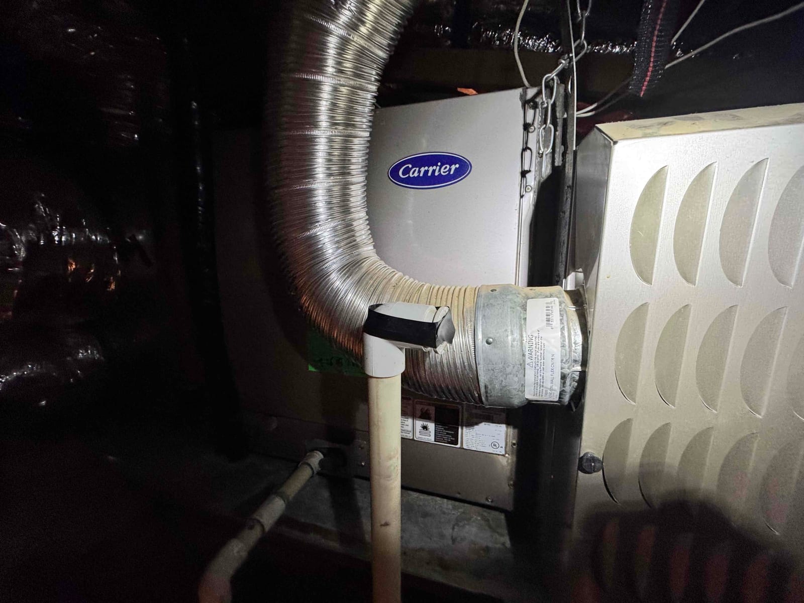 Gas furnace cabinet with flexible vent connector and PVC condensate drain line visible in the attic space.