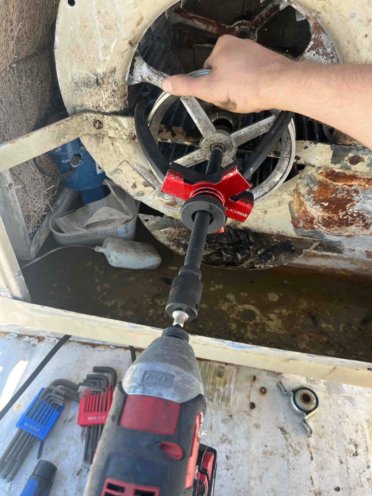 Gear puller and impact driver work on a blower wheel pulley inside a rusted housing, with hex key sets nearby.