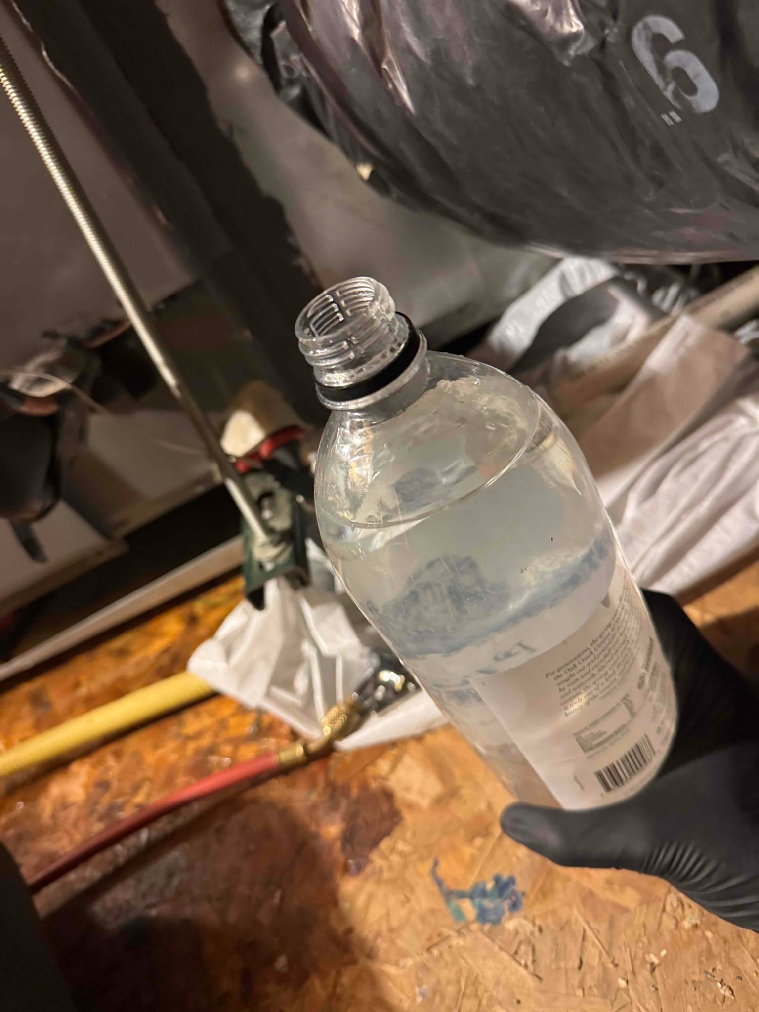 Gloved hand holds an open plastic bottle of clear liquid beside insulated ductwork, air handler cabinet, and refrigerant hoses in attic.