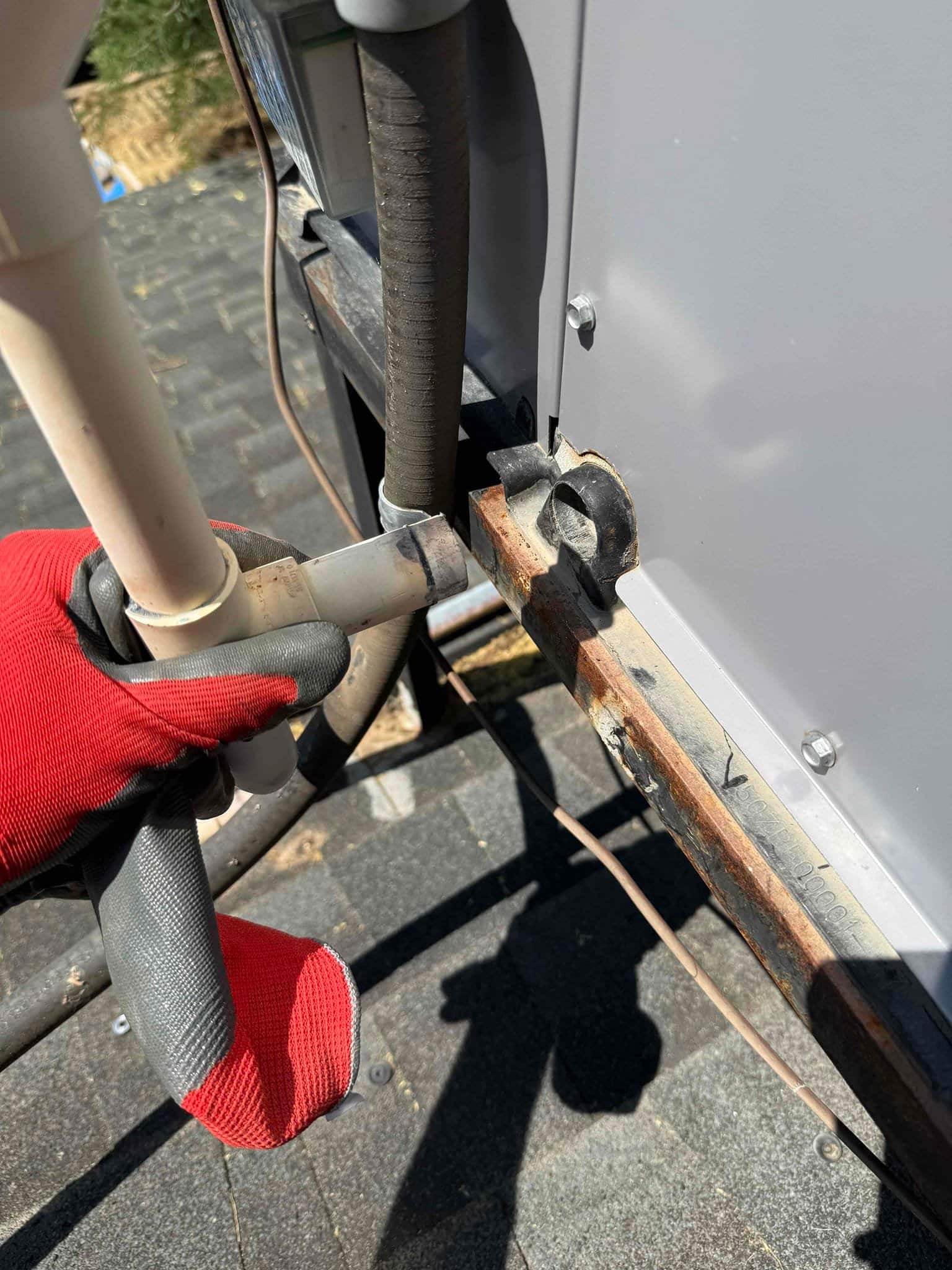 Gloved hand holds PVC condensate drain near rooftop HVAC unit, with insulated refrigerant line and rusted base rail.